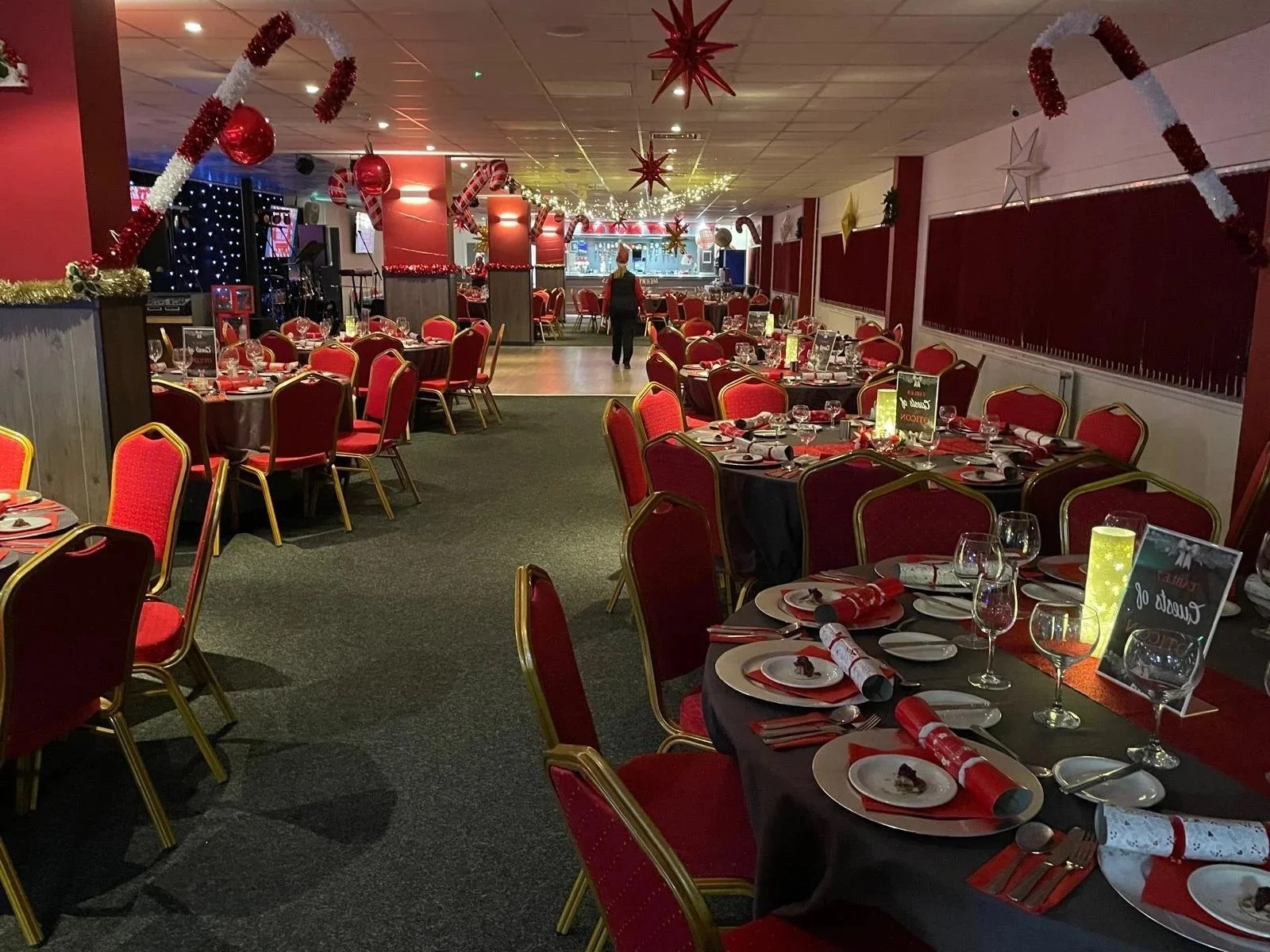 A banquet hall decorated for Christmas with red and gold decorations, tables set with plates, glasses, and Christmas-themed napkins, and a bar area in the background.