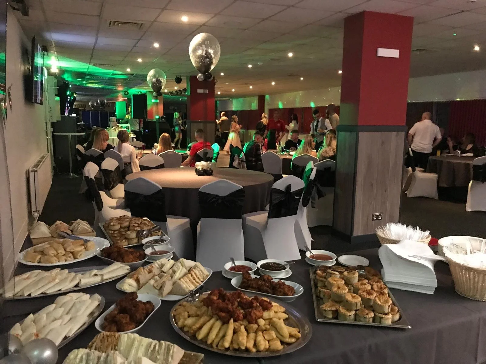 A banquet hall decorated for a celebration with tables of food, including sandwiches, snacks, and sauces, with people dining and music playing on the stage in the background. The room has black and white chairs, balloons, and green lighting.