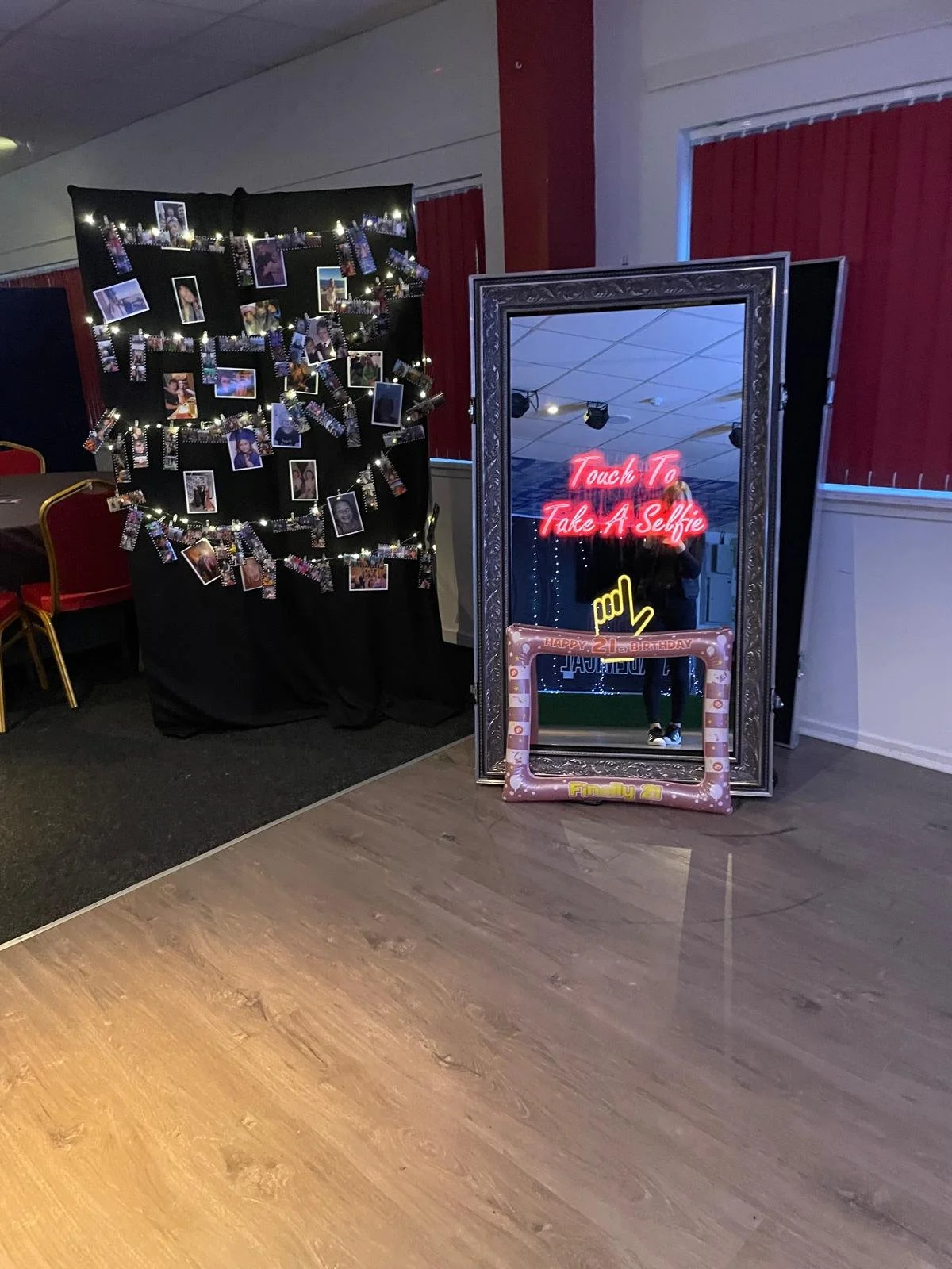 Photo of a mirror with a neon sign on it that says "Touch To Take A Selfie," and a frame below it with a message celebrating a 21st birthday. Next to the mirror is a display of photographs and decorative lights.