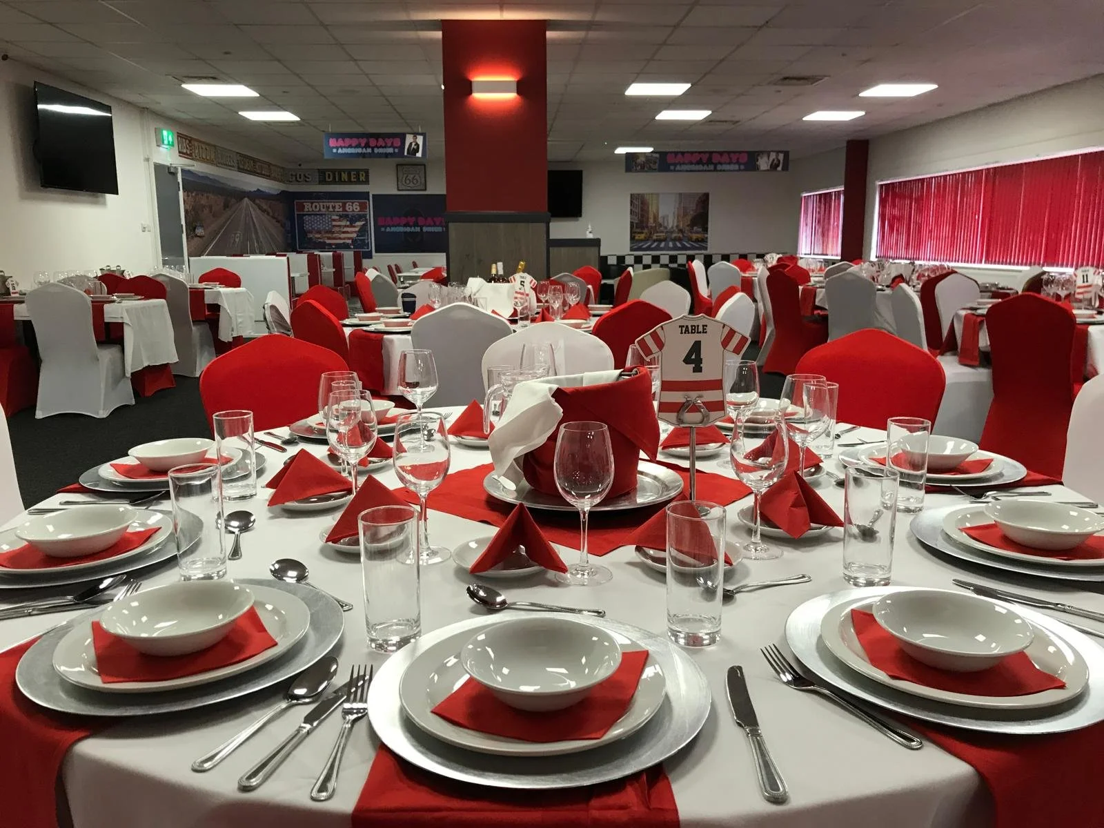 Round banquet tables set with white tablecloths, red napkins, white plates, wine glasses, water glasses, and silverware, in a decorated event space with red and white chairs, wall art, and red window blinds.