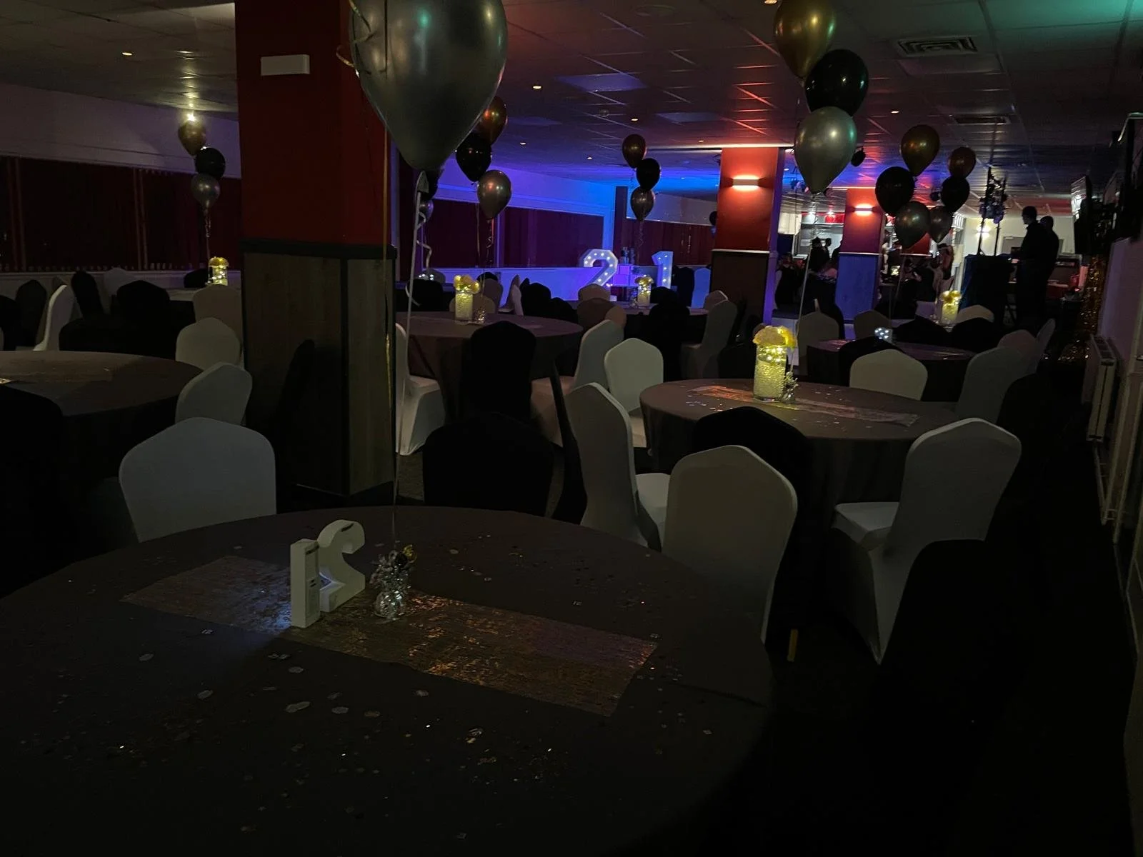 A dimly lit banquet hall decorated for a celebration with black and gold balloons, tables with black and white chairs, and a stage with illuminated numbers 2 and 1 in the background.