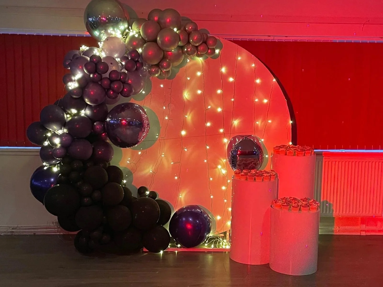 Decorative balloon arrangement with black, silver, and purple balloons illuminated by string lights, set against a red backdrop with hanging string lights, and white cylindrical pedestals with small decorations on top.