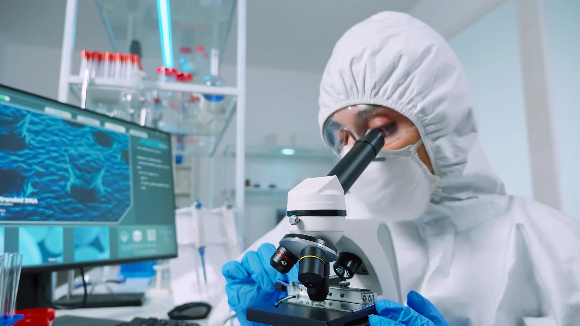 A scientist in a laboratory wearing protective gear, using a microscope and analyzing scientific data on a computer screen.