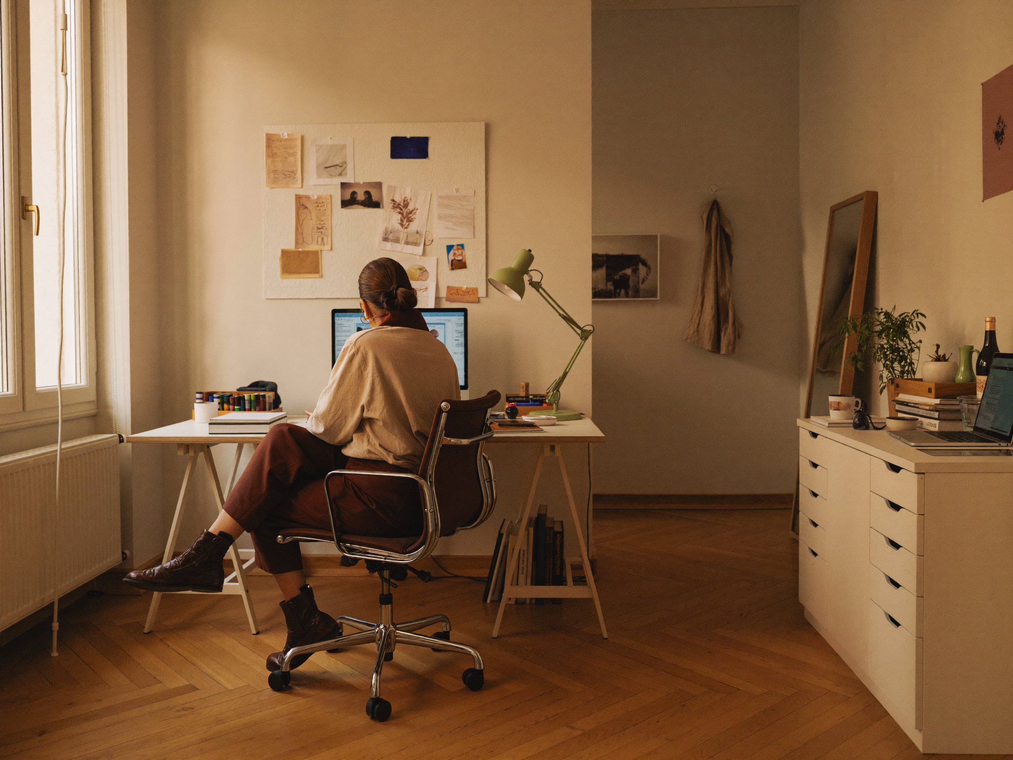A woman sits at a white desk in a cozy, well-lit room with natural light coming through a window. She is working on a computer, surrounded by various books and art supplies, with a bulletin board on the wall behind her and a large mirror leaning against the wall to the right. The room has warm wooden flooring and a cabinet with plants and personal items.
