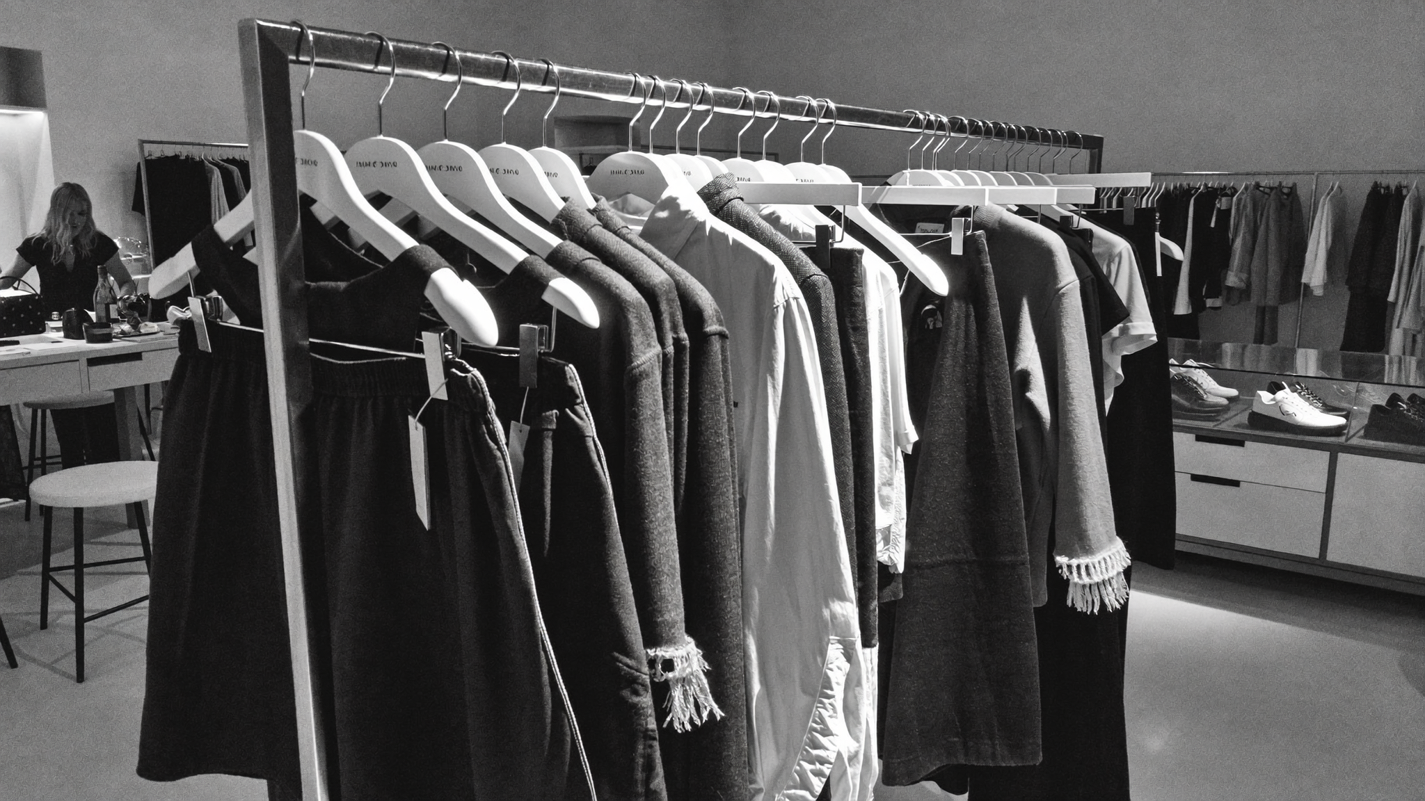 Black and white photo of clothing on display in a store, with a woman in the background near a table of accessories and shoes.