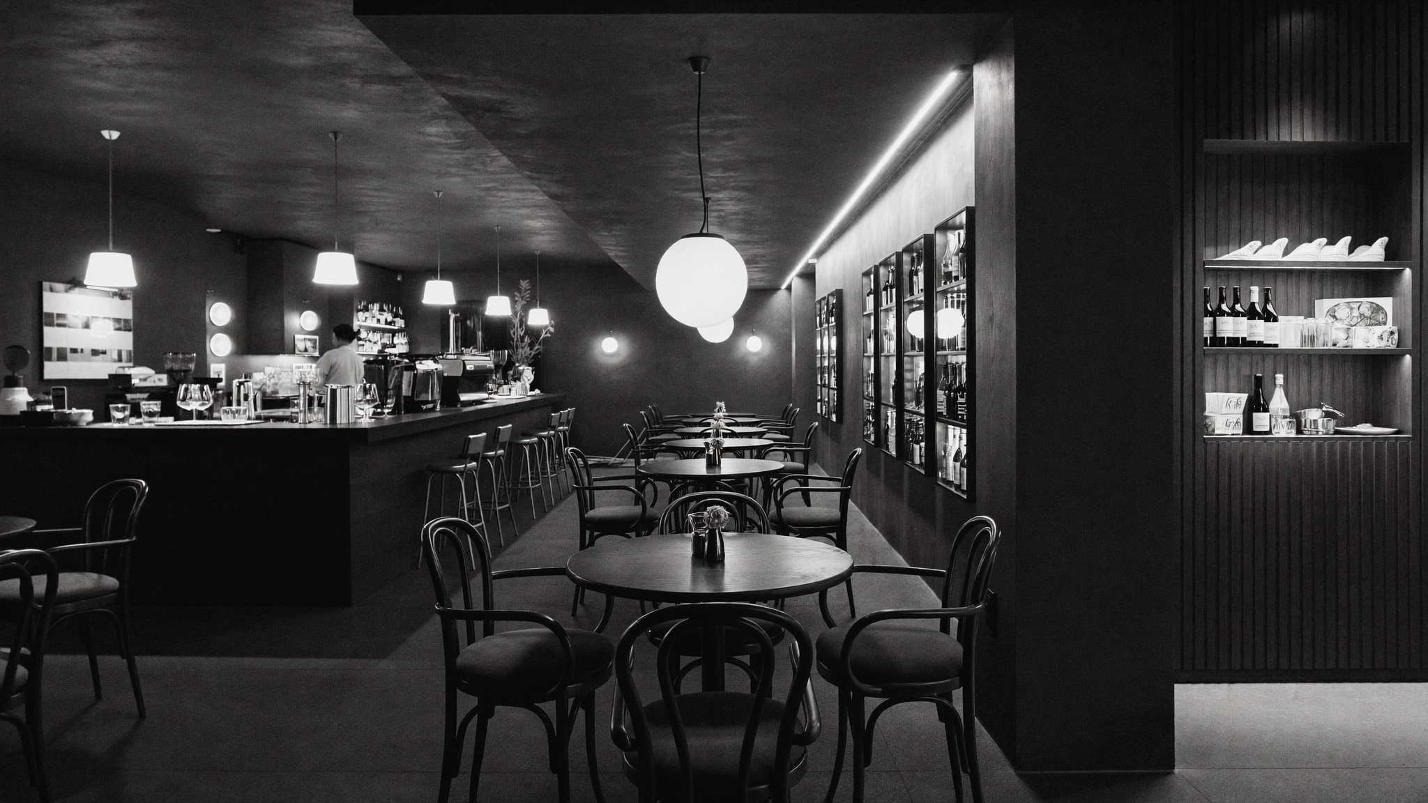 A black-and-white photo of a modern, minimalist restaurant dining area with round tables and chairs, a bar with bar stools, pendant lights, and wall-mounted shelves with bottles and decor.