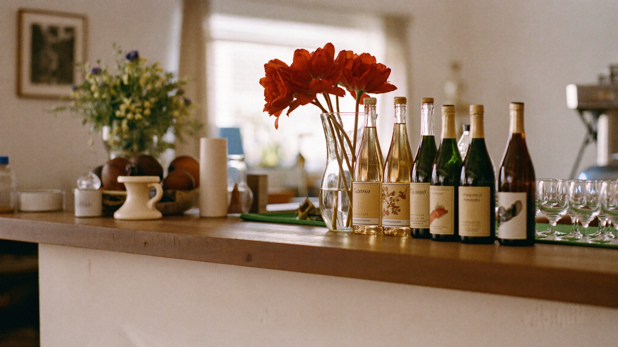 A countertop with a vase of red flowers, several bottles of wine, and empty glasses arranged in a row.