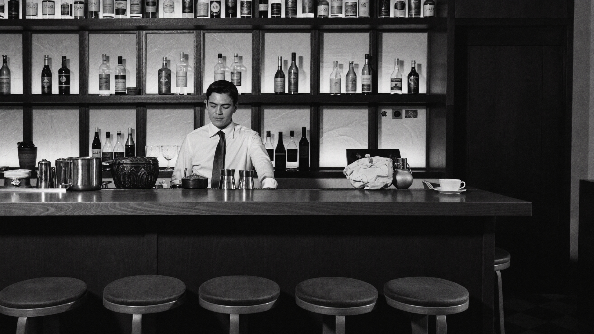 A bartender in a white shirt and tie stands behind a bar counter, with shelves of bottles in the background. The scene is in black and white.