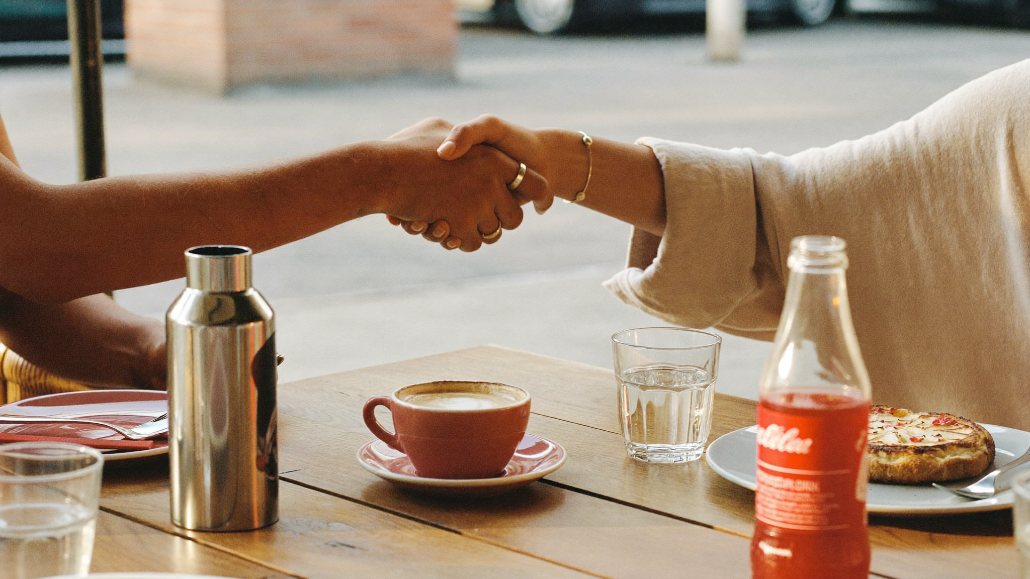 Two people shaking hands across a dining table set with drinks, a coffee cup, a glass of water, a pizza, a soda bottle, and a salt shaker.