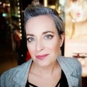 A woman with dark hair and blue eyes, wearing a gray blazer, in an indoor setting with blurry background.