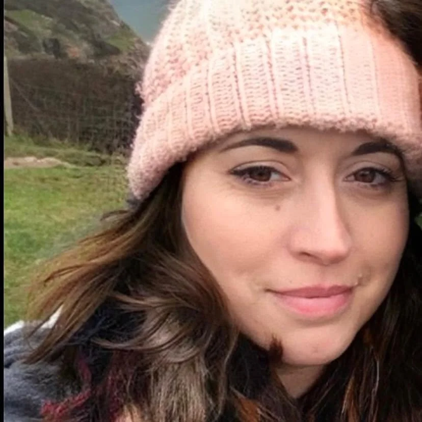 A woman wearing a pink knit beanie outdoors with a grassy hillside and trees in the background.