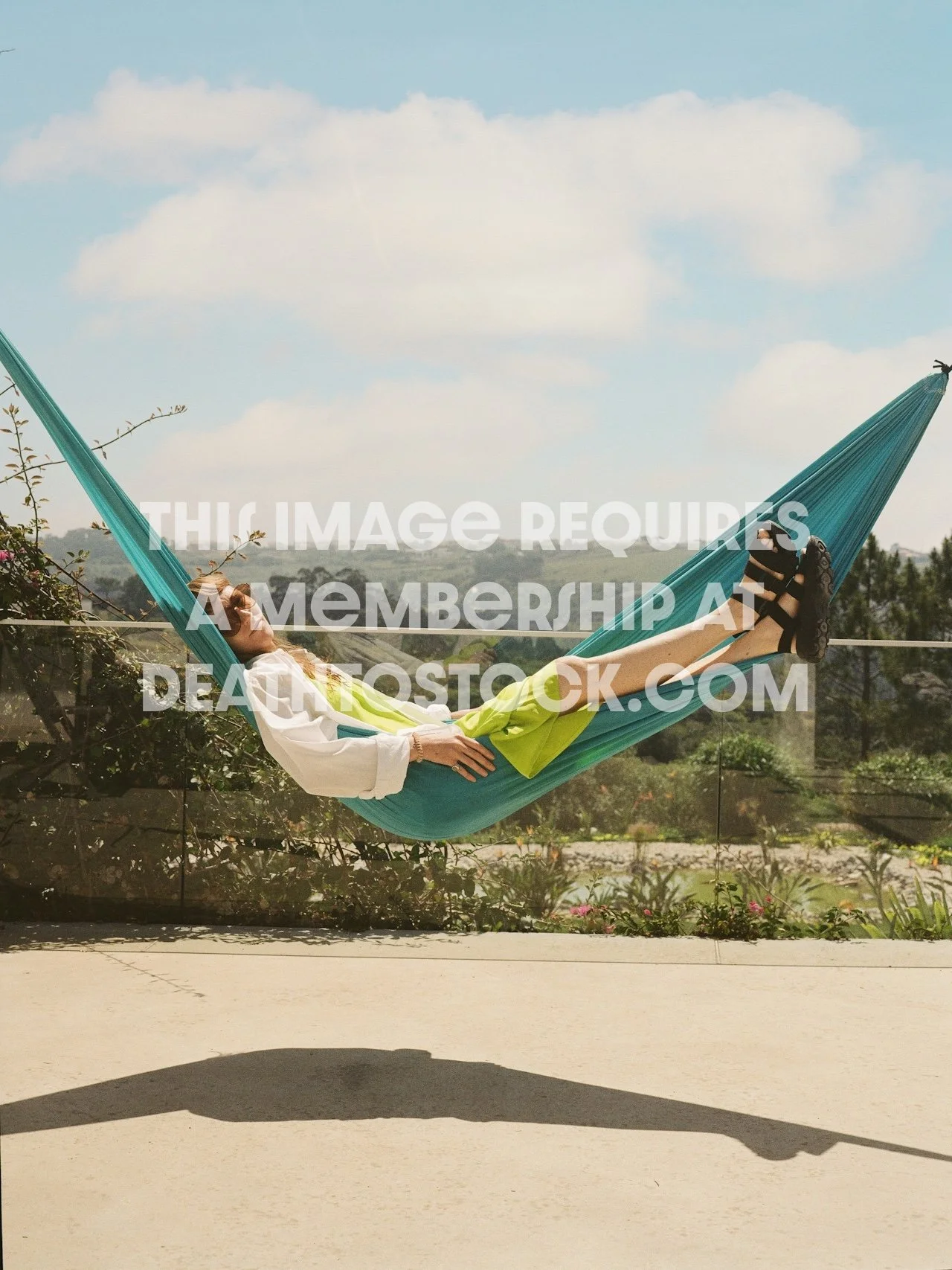 A woman lounging in a teal hammock on a sunny day, with greenery and mountains in the background.