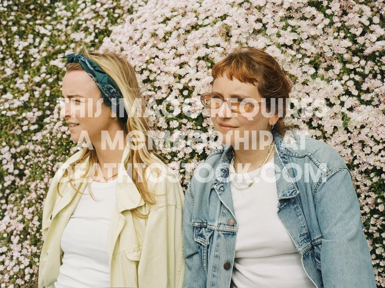 Two women standing in front of a large bush of pink and white flowers, one with blonde hair wearing a green headband and a yellow jacket, the other with short brown hair, glasses, earrings, a denim jacket, and a white shirt.