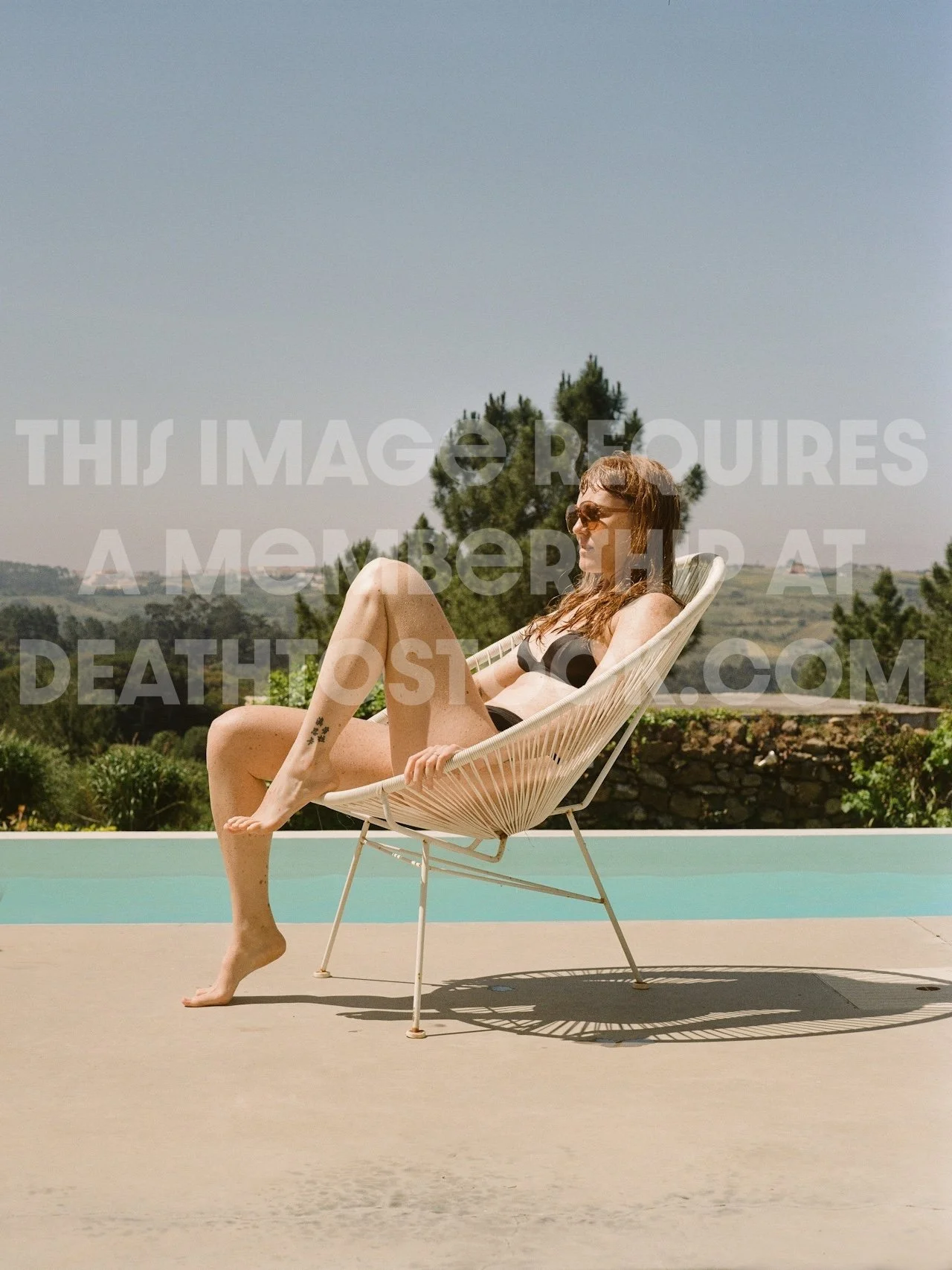 A woman in a bikini sitting on a modern white lounge chair outdoors near a pool with a scenic landscape of trees and hills in the background.