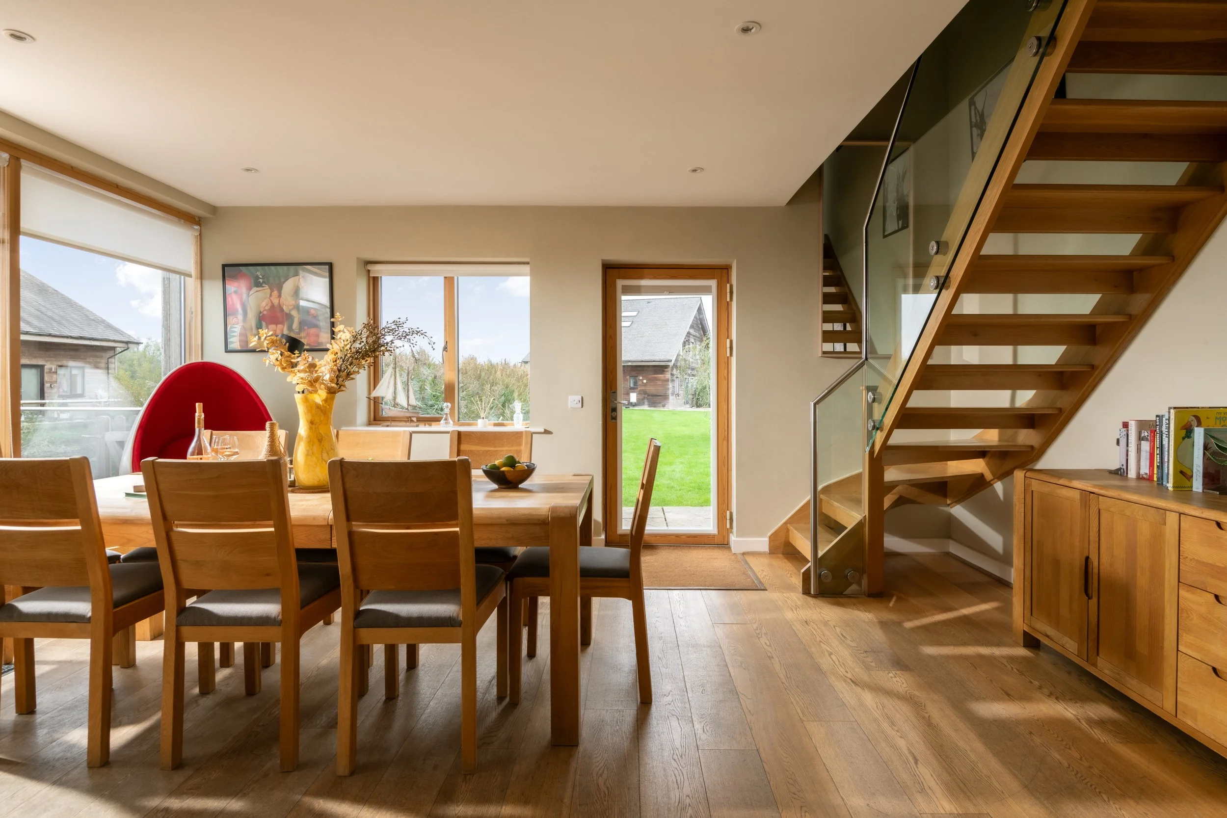 A dining area with a wooden table and chairs, large windows, a door leading outside to a grassy yard, a staircase with glass railing, a wall art, a vase with dried flowers, and a bookshelf with books.