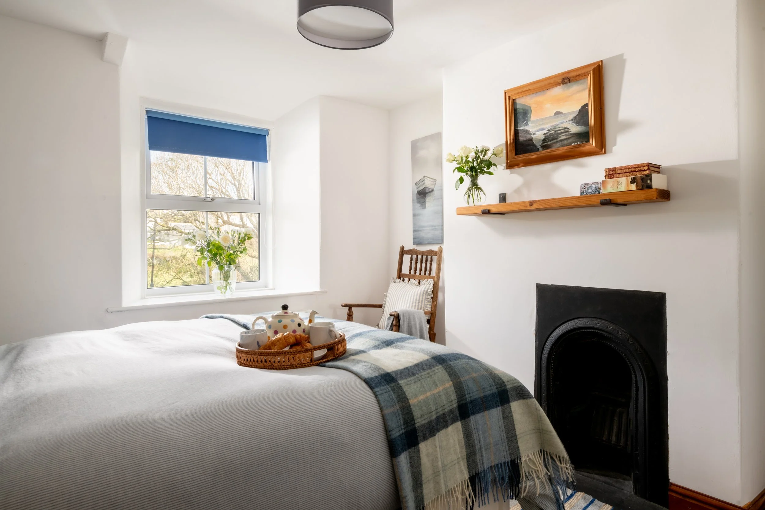 A cozy bedroom with white walls, a window with a blue shade, and a bed with a plaid blanket. On the bed, there is a wicker tray holding a teapot, two mugs, and croissants. A wooden chair with pillows and a blanket is positioned near the wall. There i