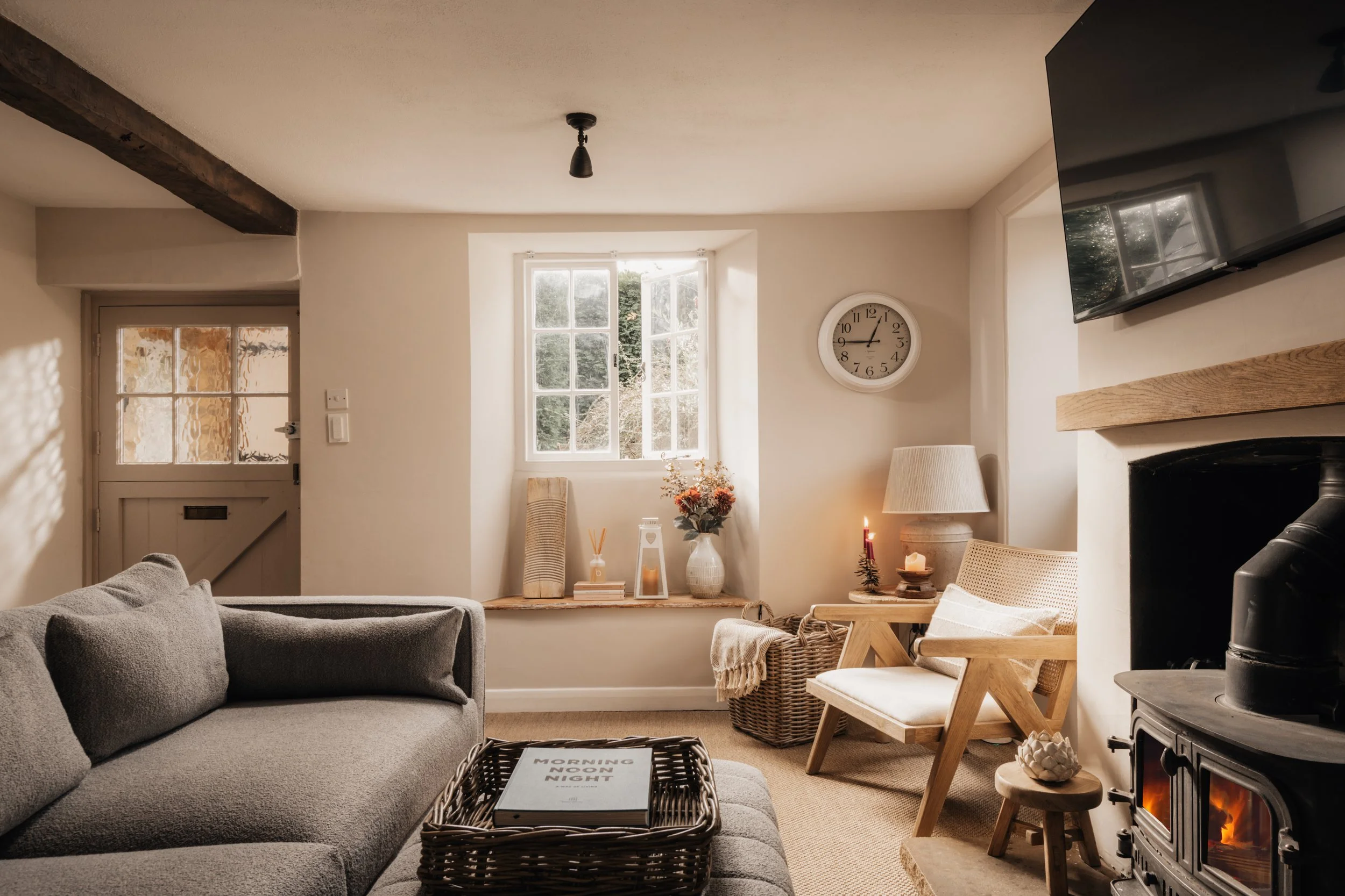 Cozy living room with gray sofa, wooden chairs, fireplace, wall clock, TV, and large windows letting in natural light.