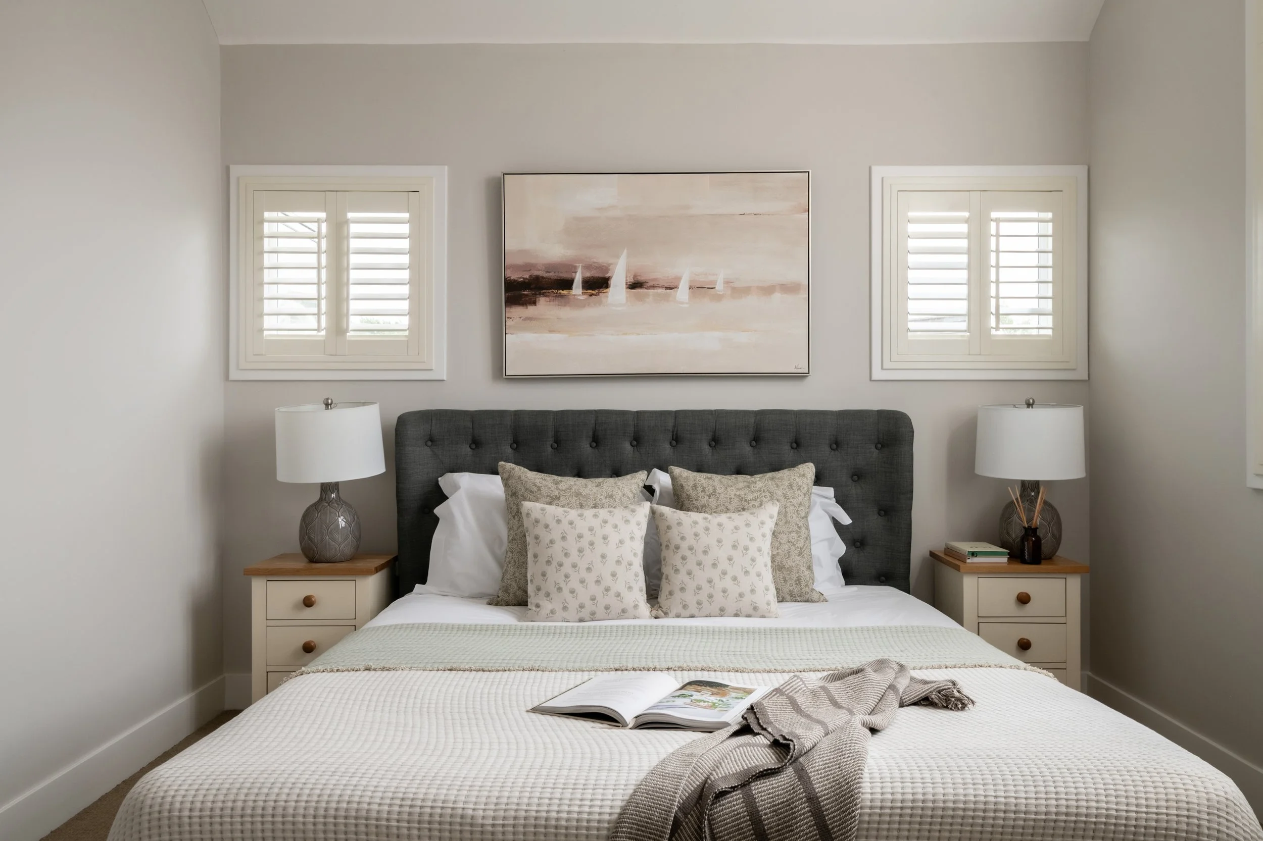 A bedroom with a large bed, two nightstands with lamps, artwork of sailboats above the bed, and two windows with shutters.