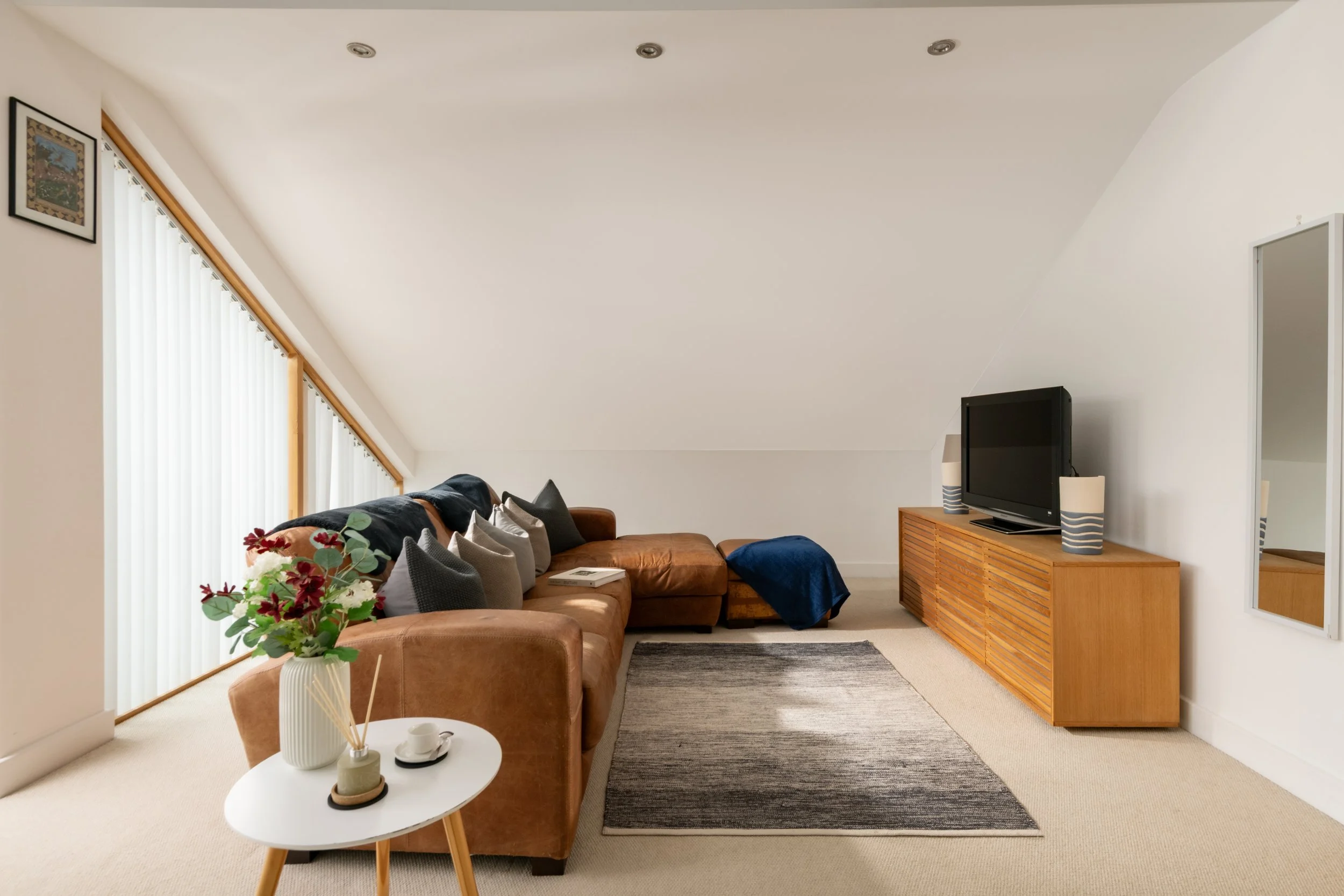 Living room with a brown sofa, colorful pillows, a white round side table with a vase of flowers, and a black TV on a wooden cabinet against a white wall with a mirror. Large windows with vertical blinds on the left side and framed artwork above. Bei