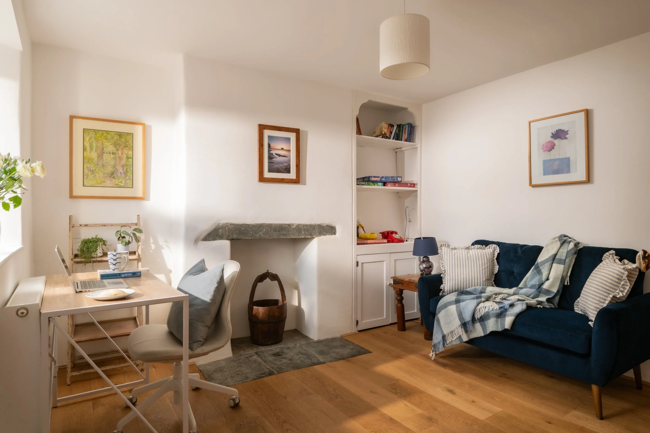 A cozy living room with white walls and hardwood floors, featuring framed artwork, a dark blue sofa with striped pillows, a white dining table with a laptop and books, a small bookshelf, and a hanging ceiling light.