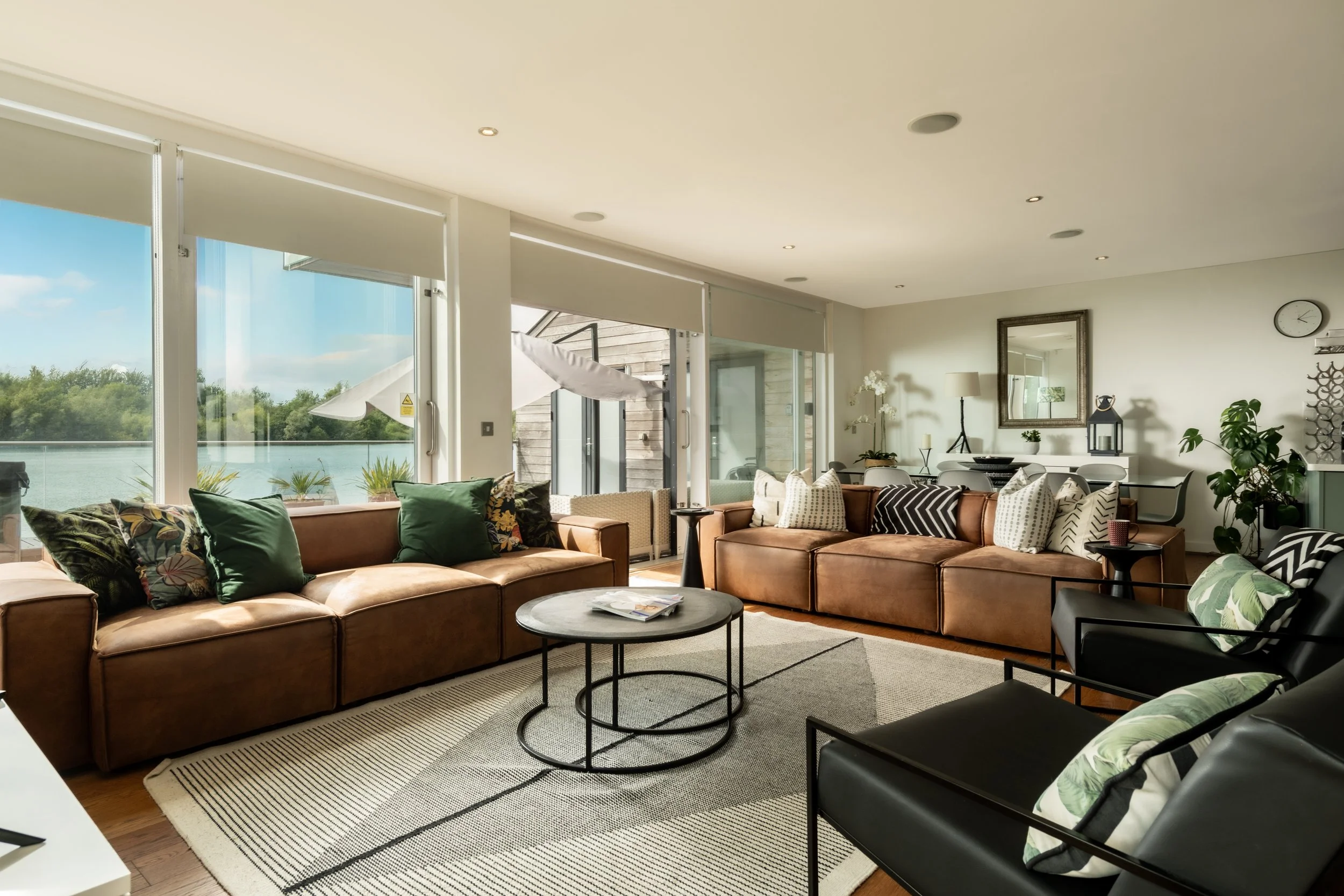 Living room with large brown leather sofas, decorated with patterned pillows, facing a view of water through large windows, with indoor plants and wall decor.