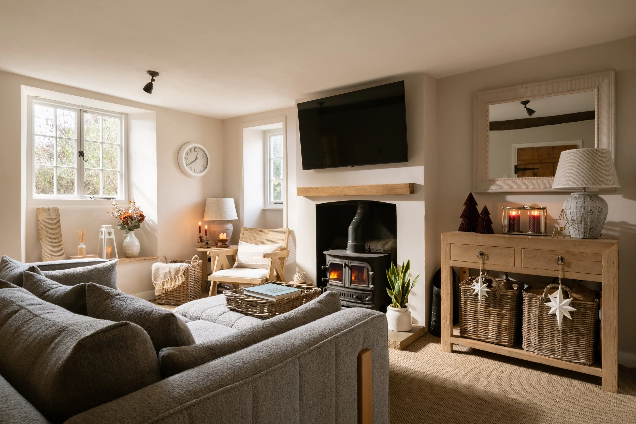 A cozy living room with beige walls and natural light from two large windows, decorated with a clock, candles, plants, and holiday ornaments, with a wood-burning stove and mounted TV over a fireplace.