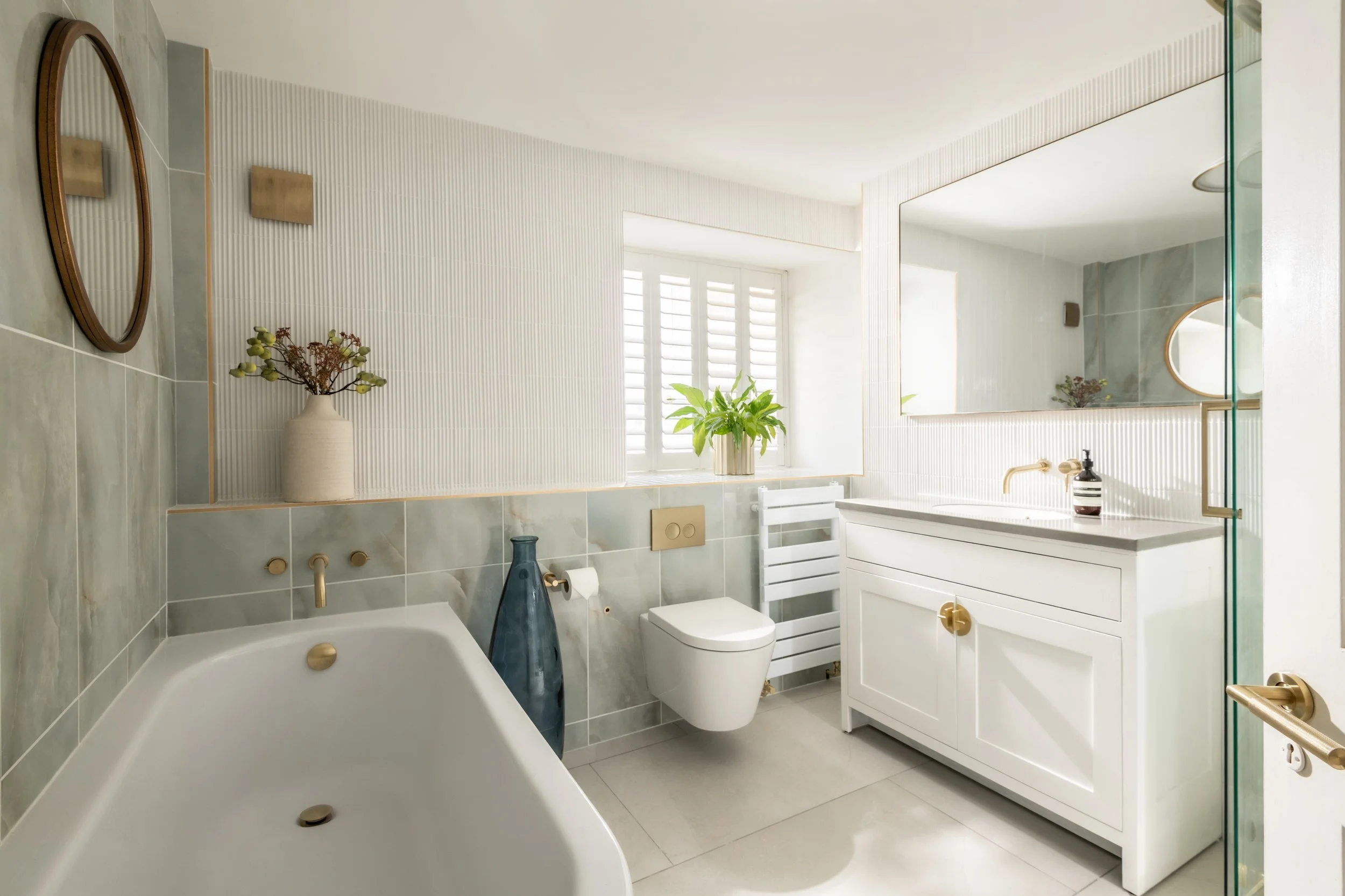 Modern bathroom with a bathtub, a white toilet, and a white vanity with a mirror above it. Decor includes vases with flowers and plants, and a window with white shutters.