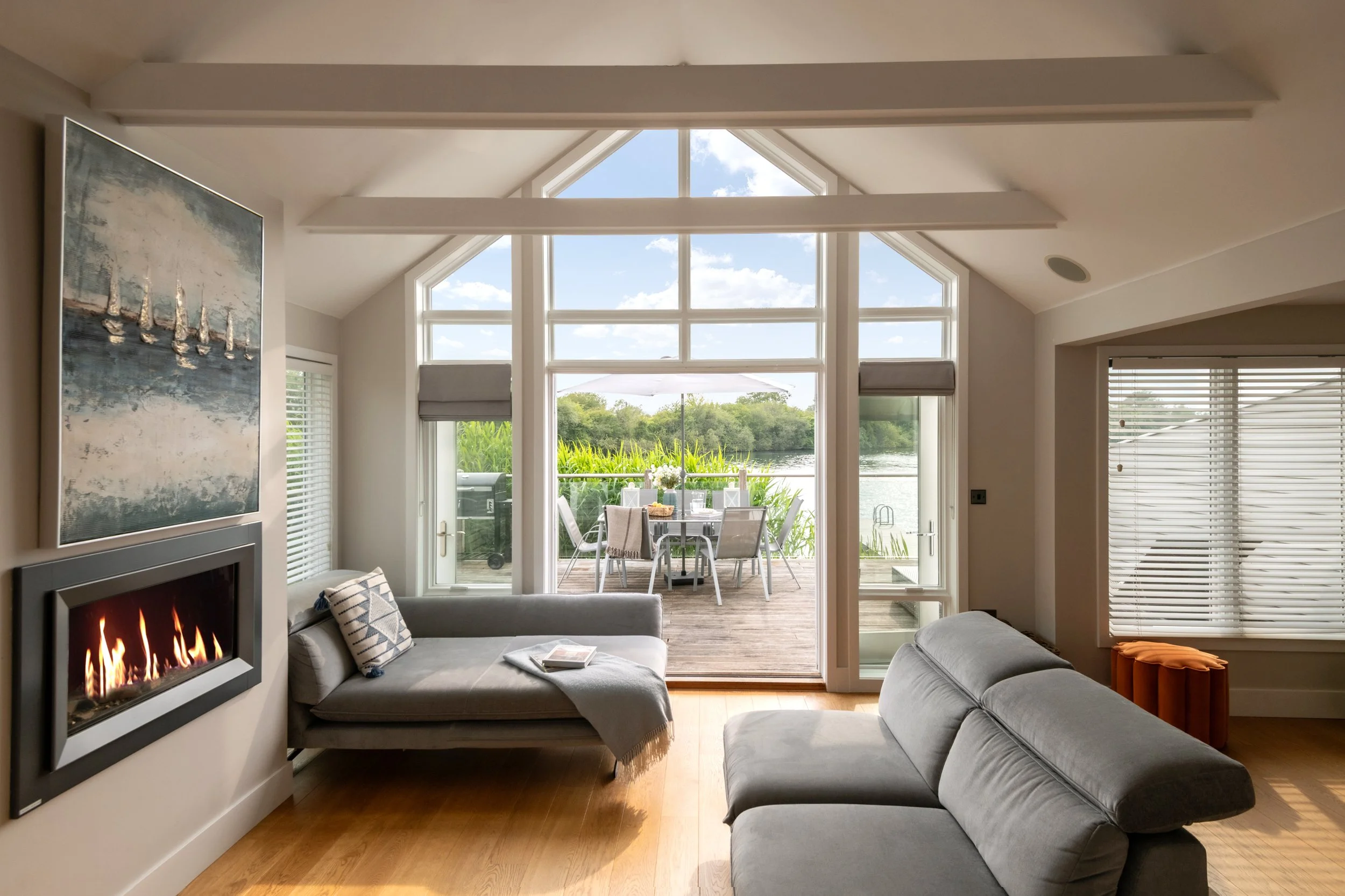 Living room with gray sofas, a fireplace, large windows with a view of a deck and water, indoor plants, and outdoor table with chairs.