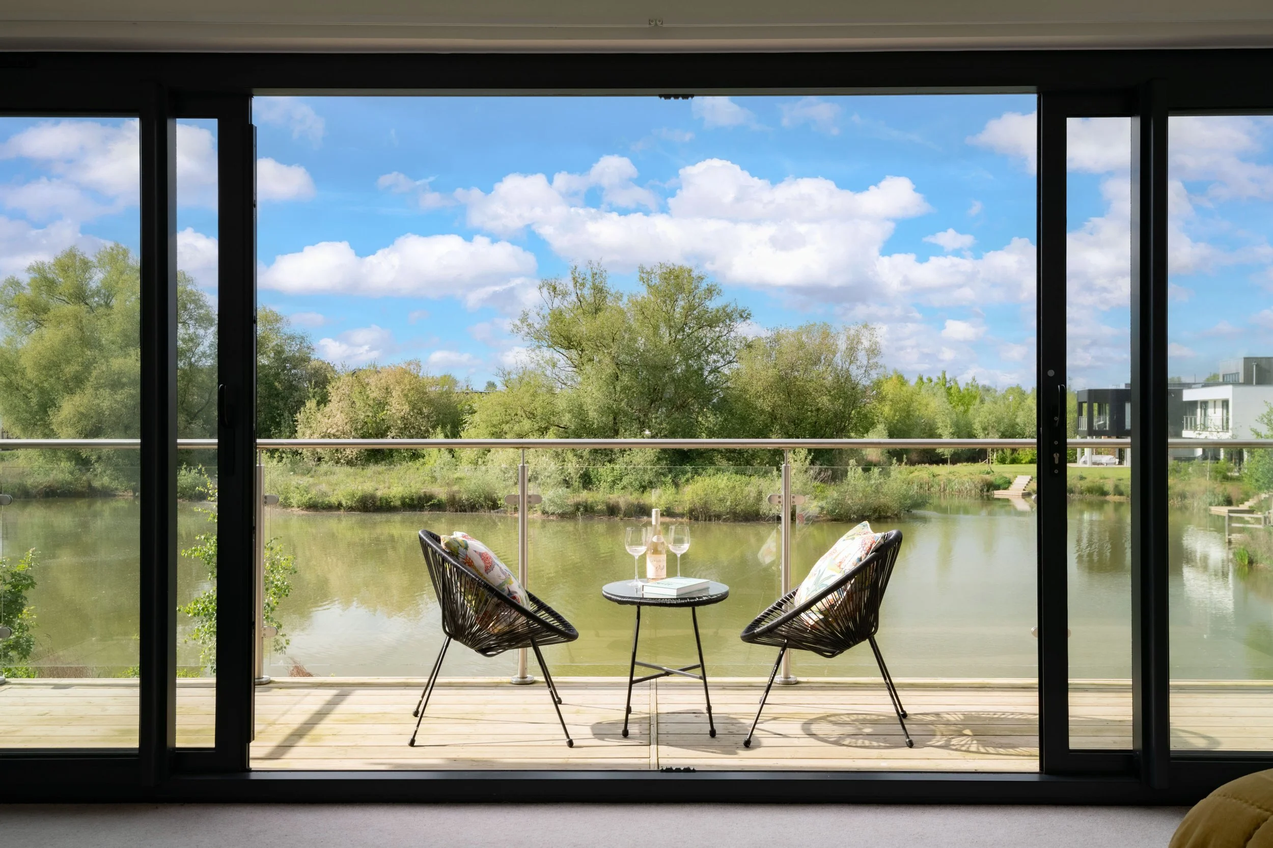 View from a room showing a balcony with two chairs and a small table holding two glasses and a bottle, overlooking a river, lush green trees, and modern houses under a partly cloudy sky.