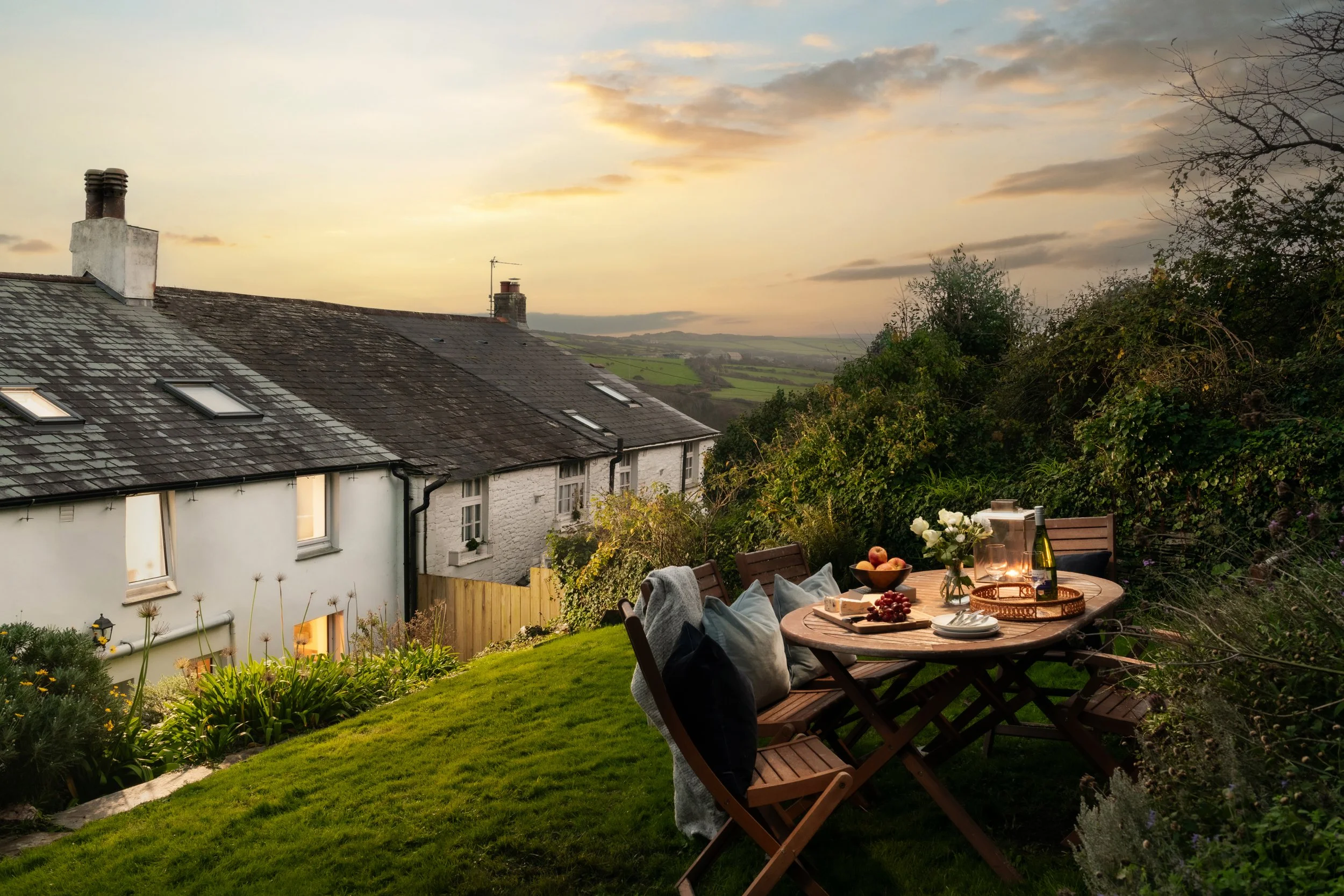 An outdoor patio table set for a meal with wine, apples, and bread, surrounded by chairs and greenery, overlooking rooftops and a countryside landscape during sunset.