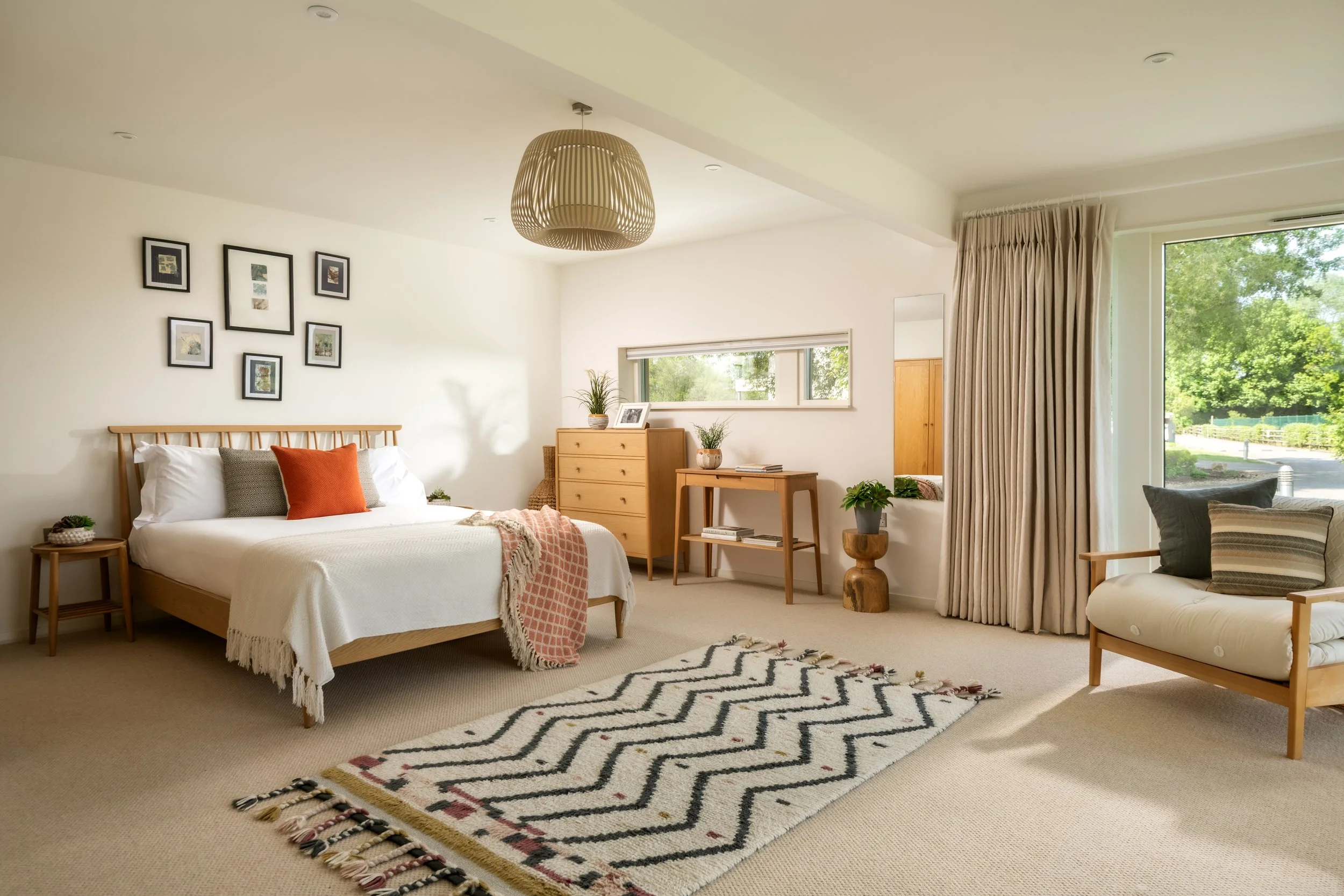 A bedroom with a wooden bed frame, white bedding, and decorative pillows, a wooden dresser, a small side table, and colorful rugs on a beige carpet, with large windows and light curtains, and a modern ceiling light.
