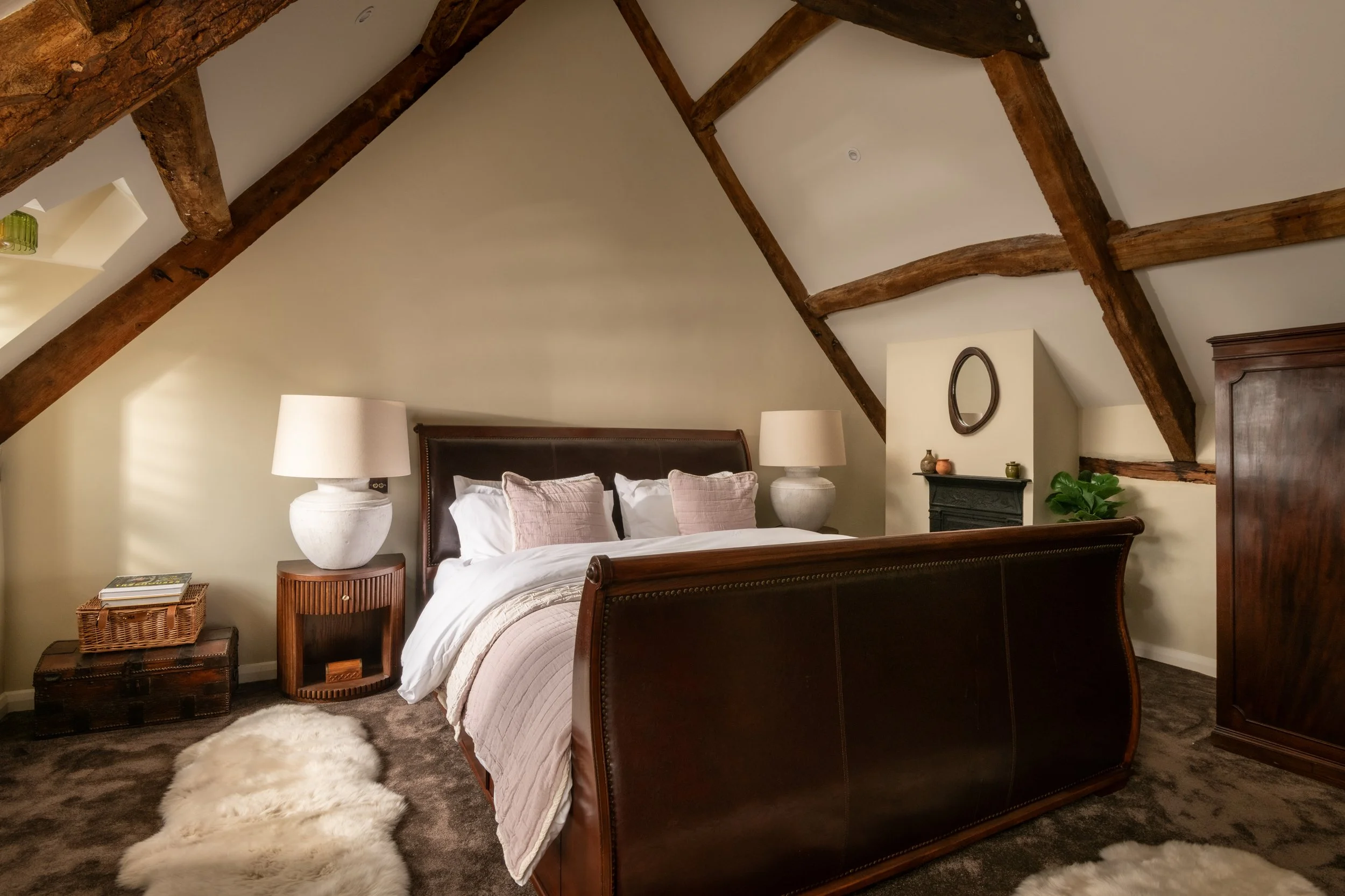 A bedroom with a dark wooden bed frame, white bedding, and pillows. Flanking the bed are matching white lamps on round wooden nightstands. There is a woven basket with books on it on the left and a cream-colored furry rug on the dark carpet in front 
