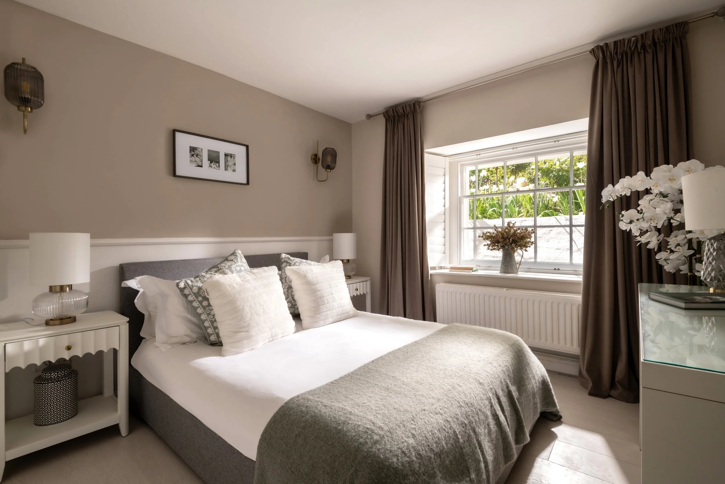 A cozy bedroom with a large window letting in natural light, decorated with neutral tones, white bedside tables, and lamps, along with framed pictures on the wall and a vase with pink flowers on the windowsill.