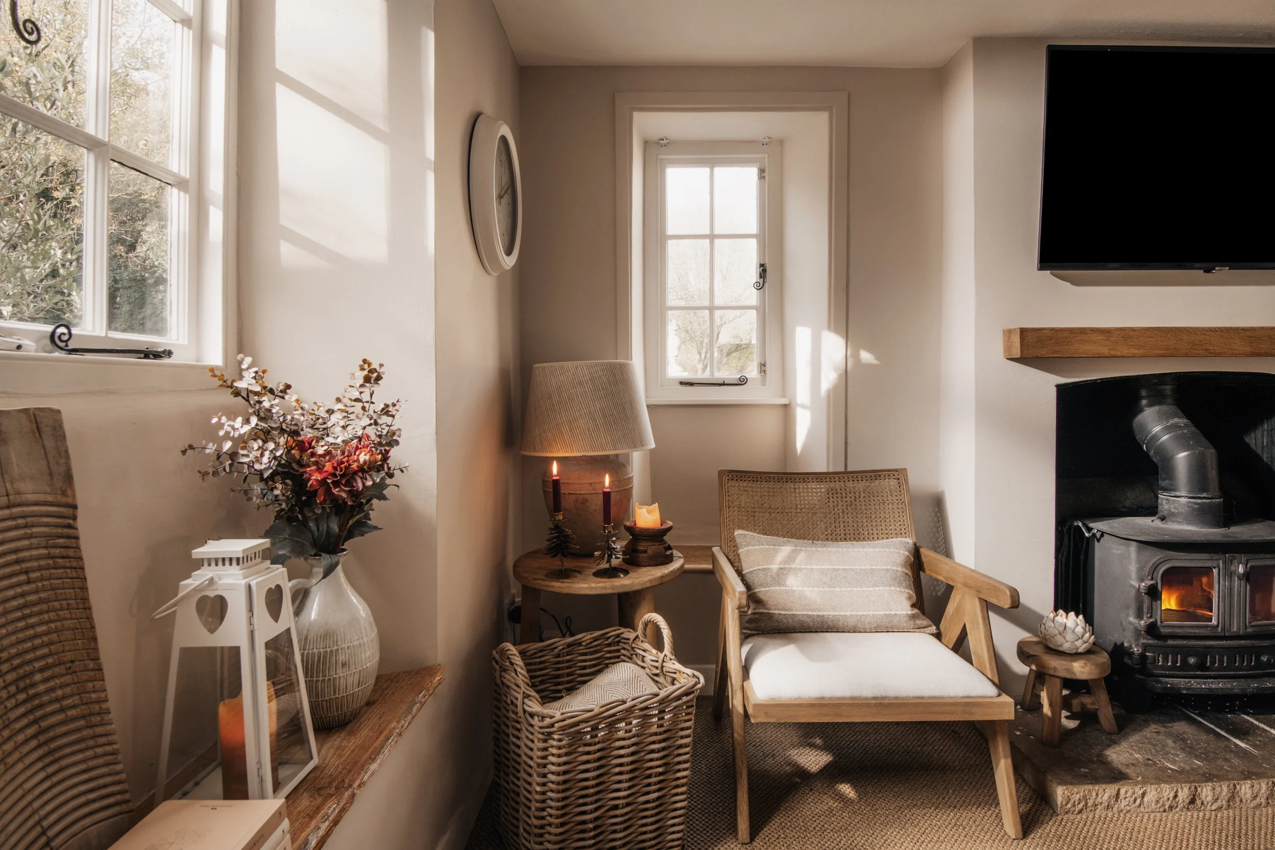 Sunlit cozy corner with two windows, a wood-burning stove, a beige armchair, wicker basket, flowering plant, candles, and decorative items.