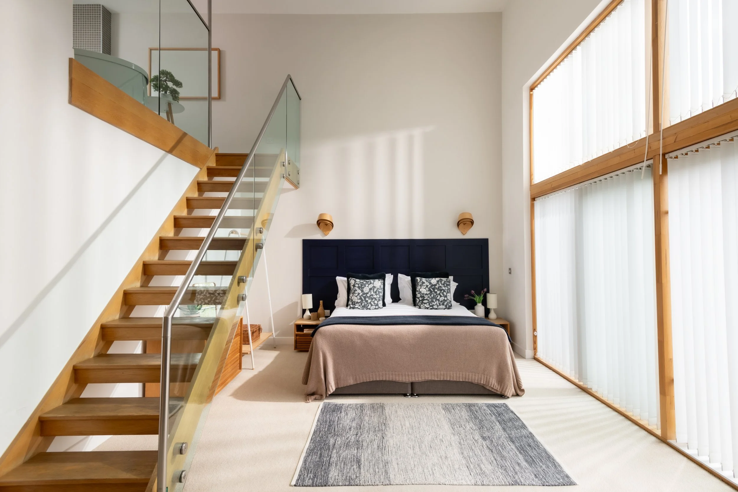 Modern bedroom with high ceilings, large windows with vertical blinds, a bed with patterned pillows, nightstands with lamps, and a wooden staircase with glass railings.