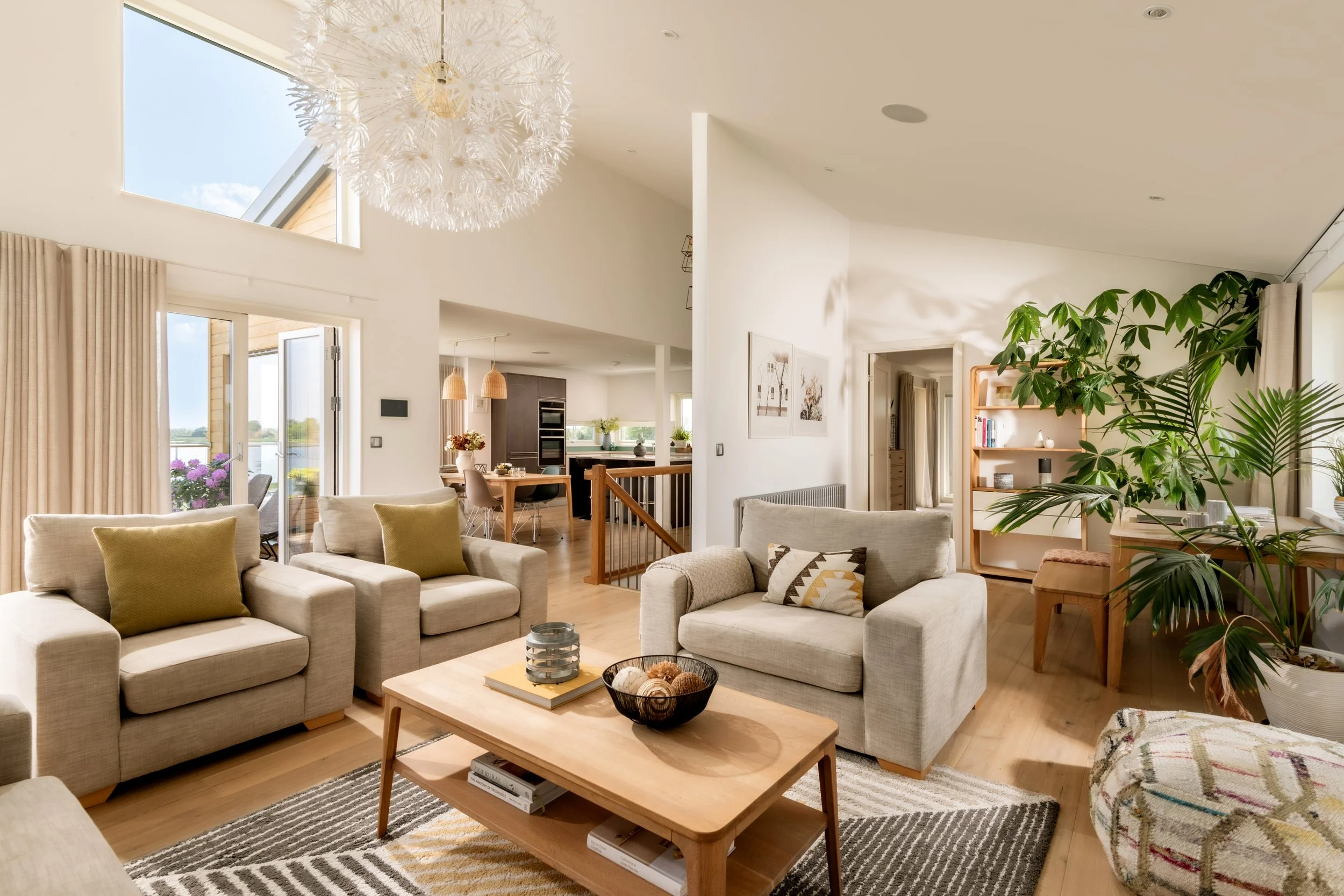 Bright and airy living room with beige sofas, a wooden coffee table, large windows, a chandelier, and indoor plants.