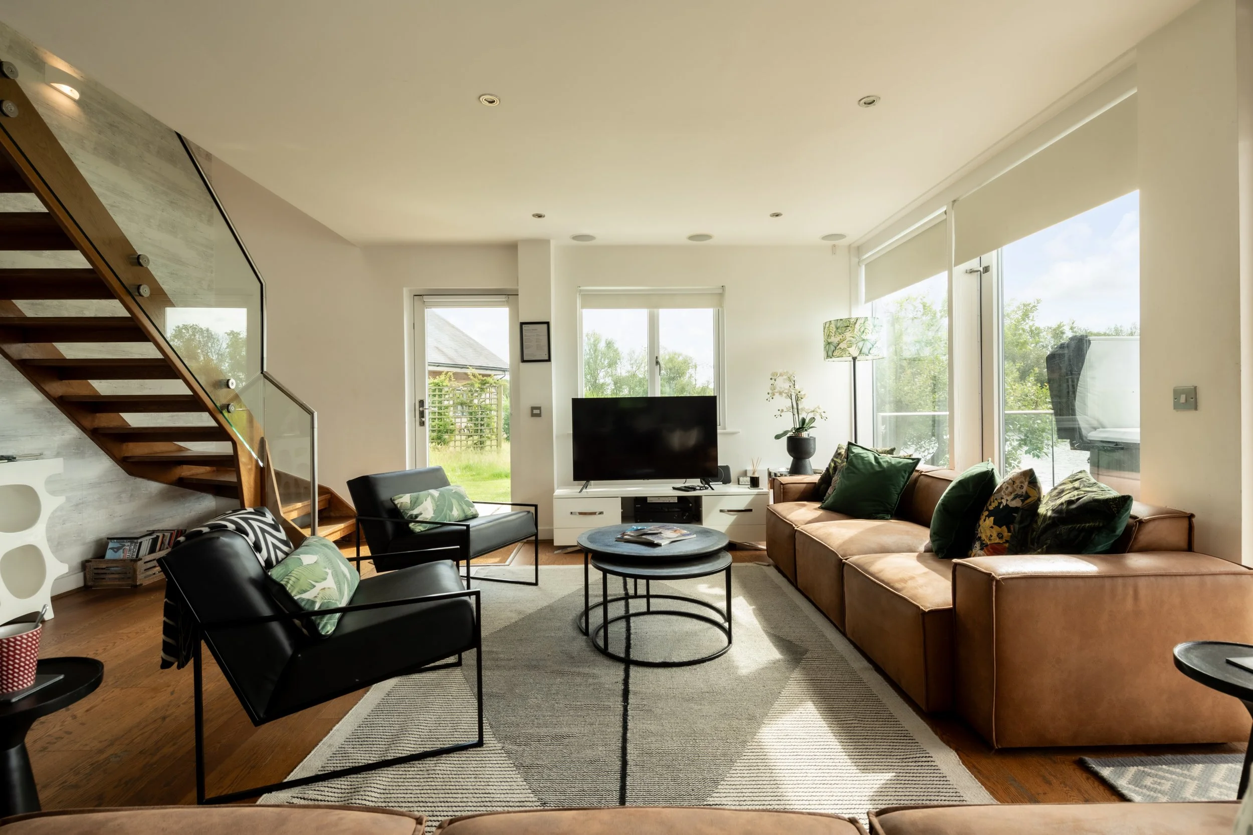 Modern living room with tan sofa, black chairs, and coffee tables, large windows with blinds, and a staircase on the left, decorated with plants and accessories.