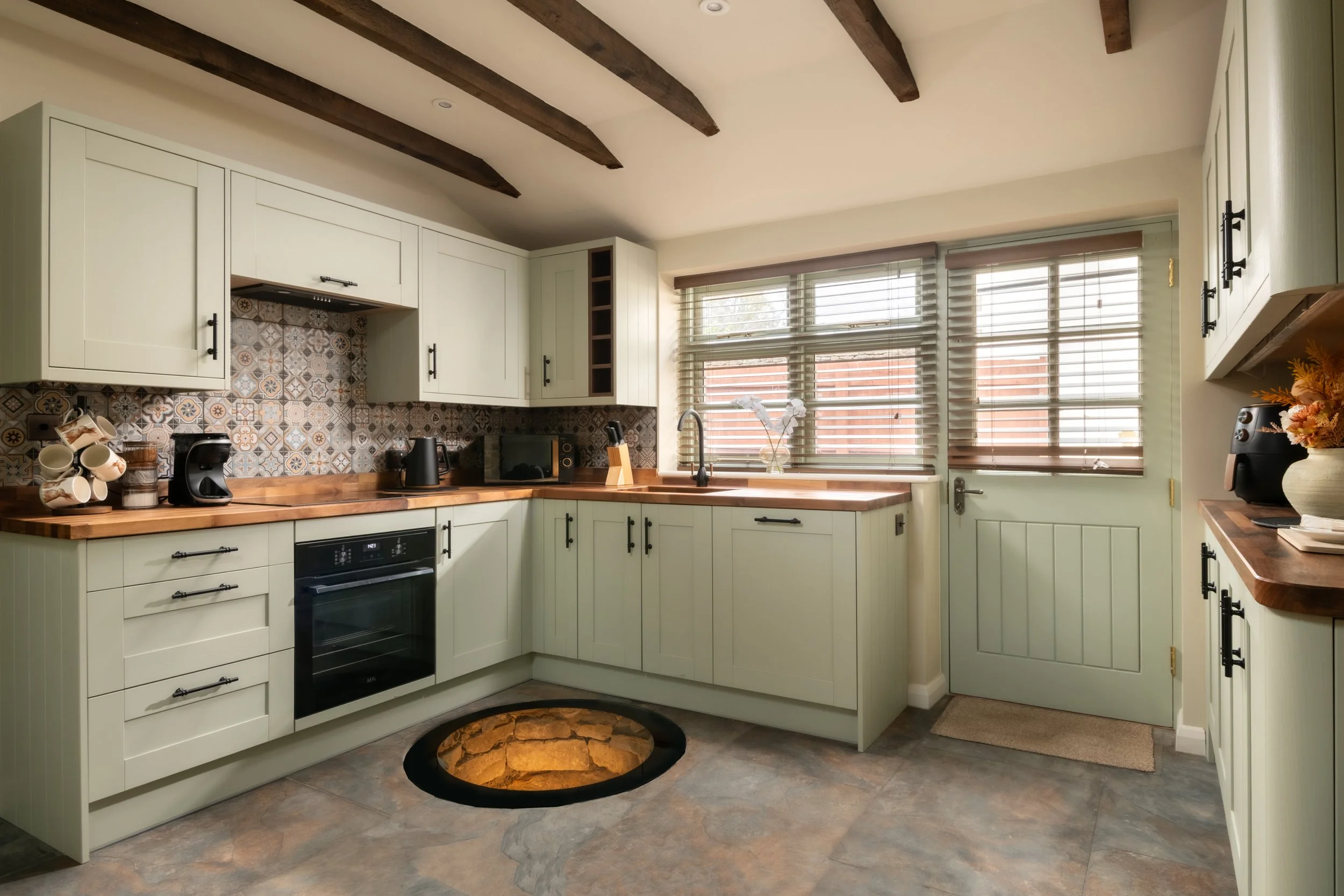 A kitchen with cream-colored cabinets, wooden countertops, and a patterned tile backsplash. It features a black oven, coffee maker, microwave, and a sink with a faucet. Large windows with blinds allow natural light, and a flower vase is on the window