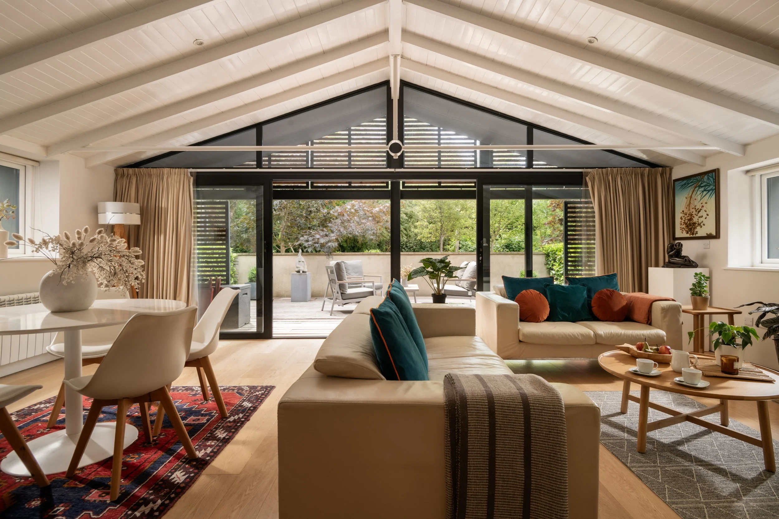 A spacious living room with a vaulted ceiling, large glass doors leading to an outdoor patio with chairs, and various seating with colorful pillows. There are plants, framed artwork, and a side table with decorative items.