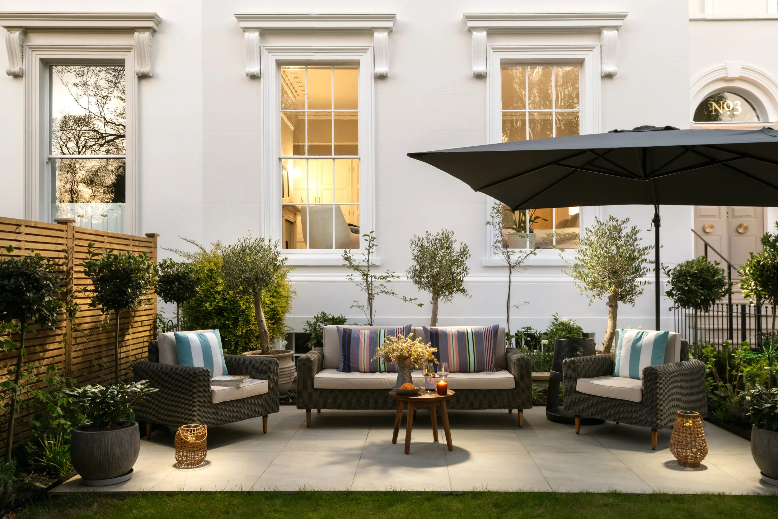 A backyard patio with outdoor furniture including a sofa and chairs with striped cushions, a small wooden table with candles, potted plants, a large patio umbrella, and a white house with tall windows in the background.
