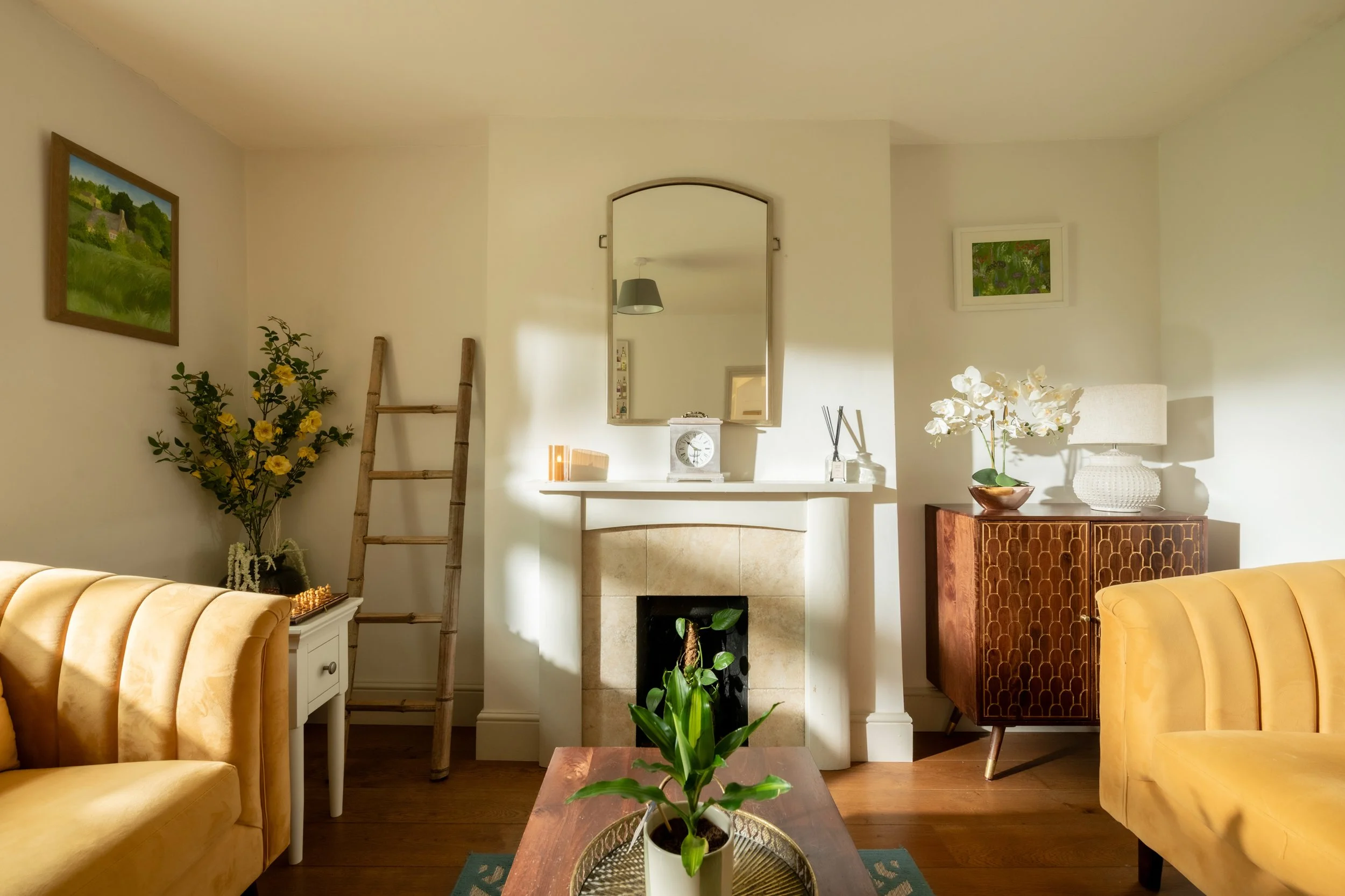 Living room with yellow velvet armchairs, a white side table, a wooden coffee table with a potted plant, a fireplace with a mirror above, and wall art. Decor includes a tall plant with yellow flowers, a lamp, and decorative items.
