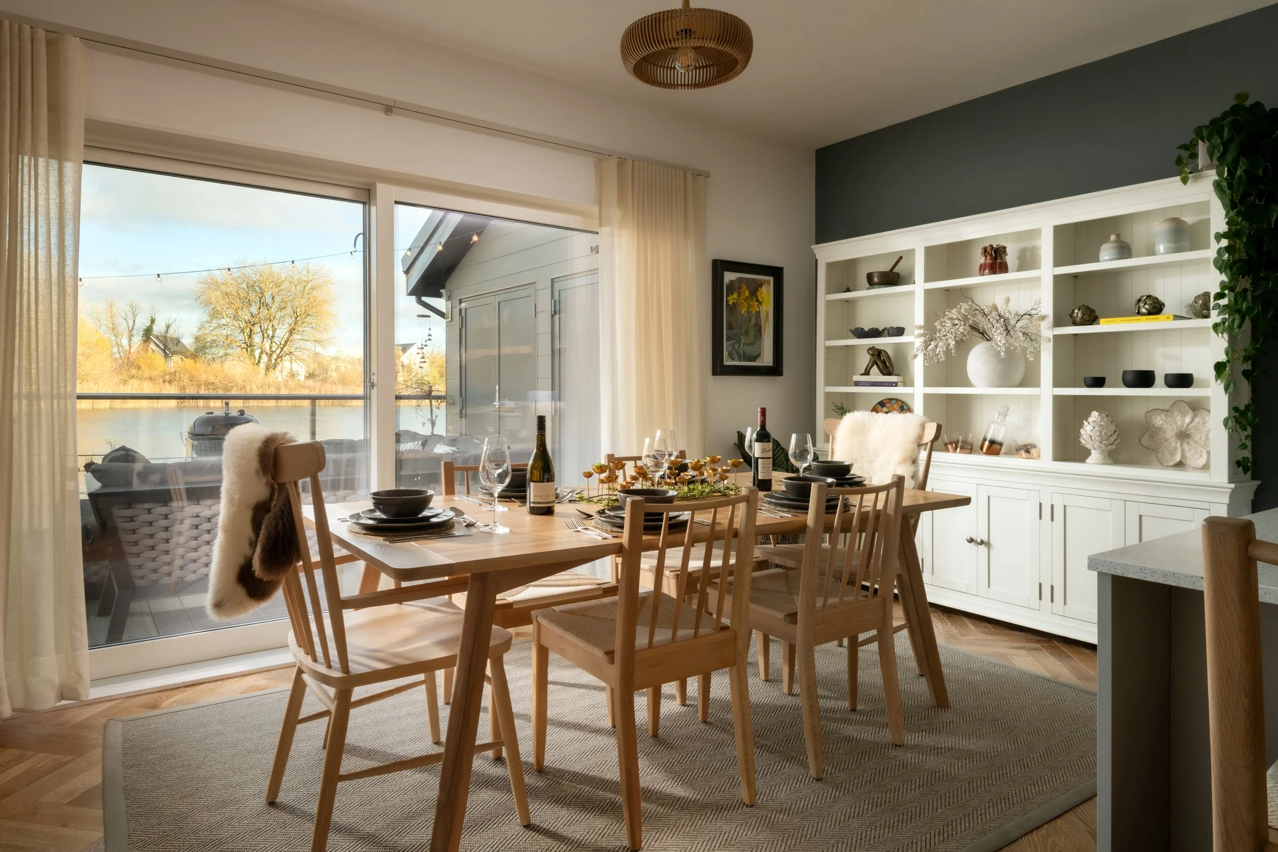 Dining room with a large wooden table set with wine glasses, black bowls, a bottle of wine, and a decorative centerpiece. There are wooden chairs, some with furry throws. A large window with sheer curtains offers a view of trees and a body of water o