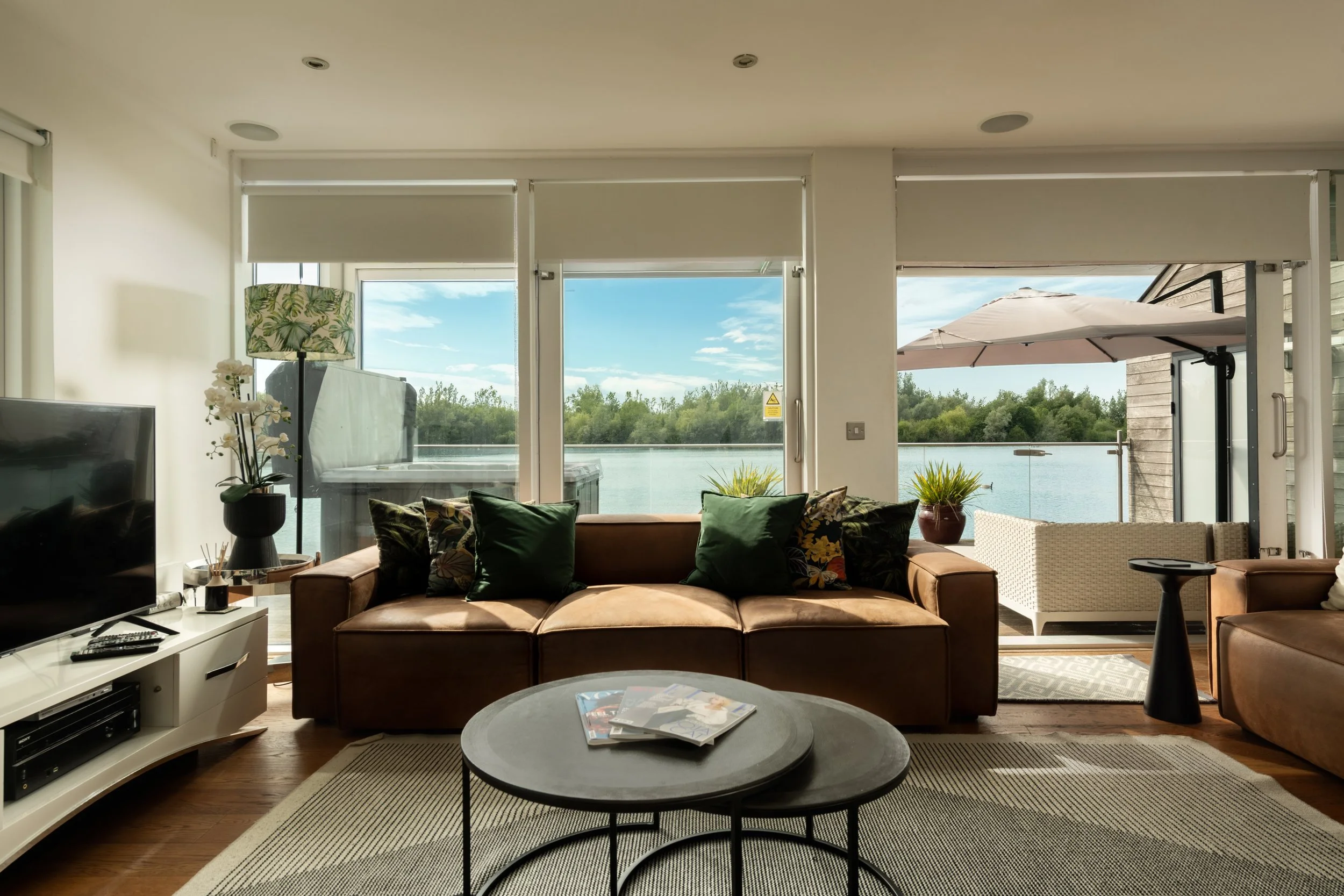 Living room with large windows overlooking a lake, tropical decor, brown sofa with green and floral pillows, coffee table with magazines, white TV stand, and outdoor patio with umbrella.