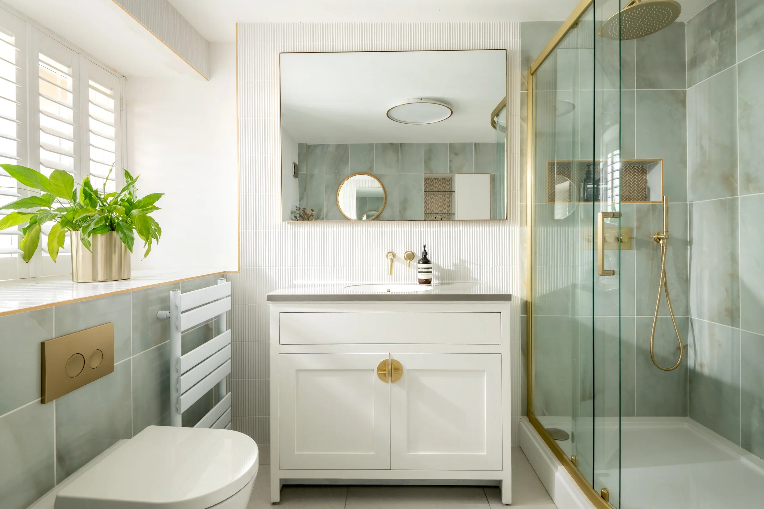 Modern bathroom with light gray tiled walls, white vanity with gold accents, a large mirror, a potted green plant sitting on a window sill, and a glass-enclosed shower with a gold showerhead.