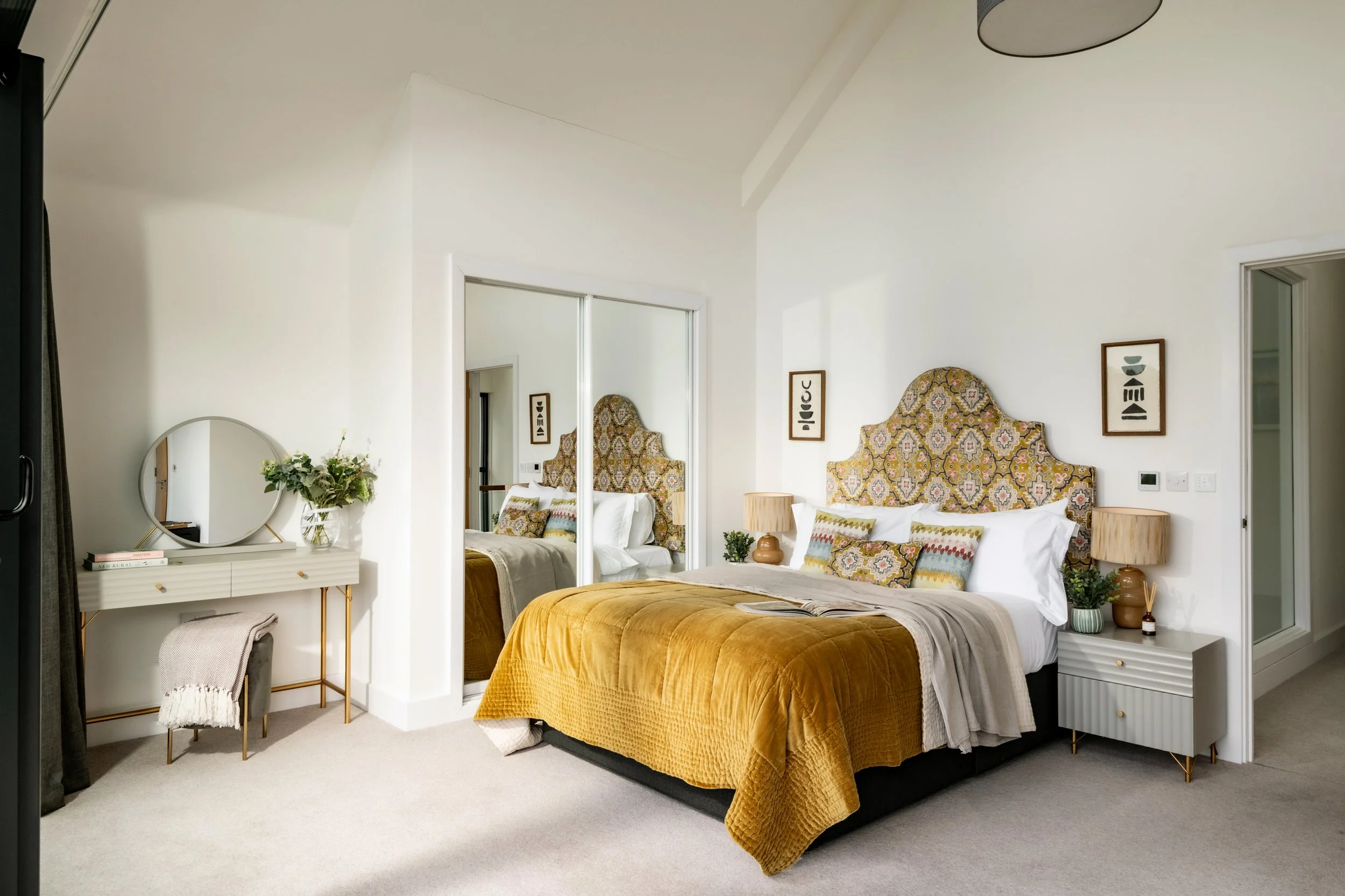 A neatly made bed with a mustard yellow quilt, patterned pillows, and a decorative headboard in a modern bedroom. There are two bedside tables with lamps, a mirror, and framed artwork on the walls.