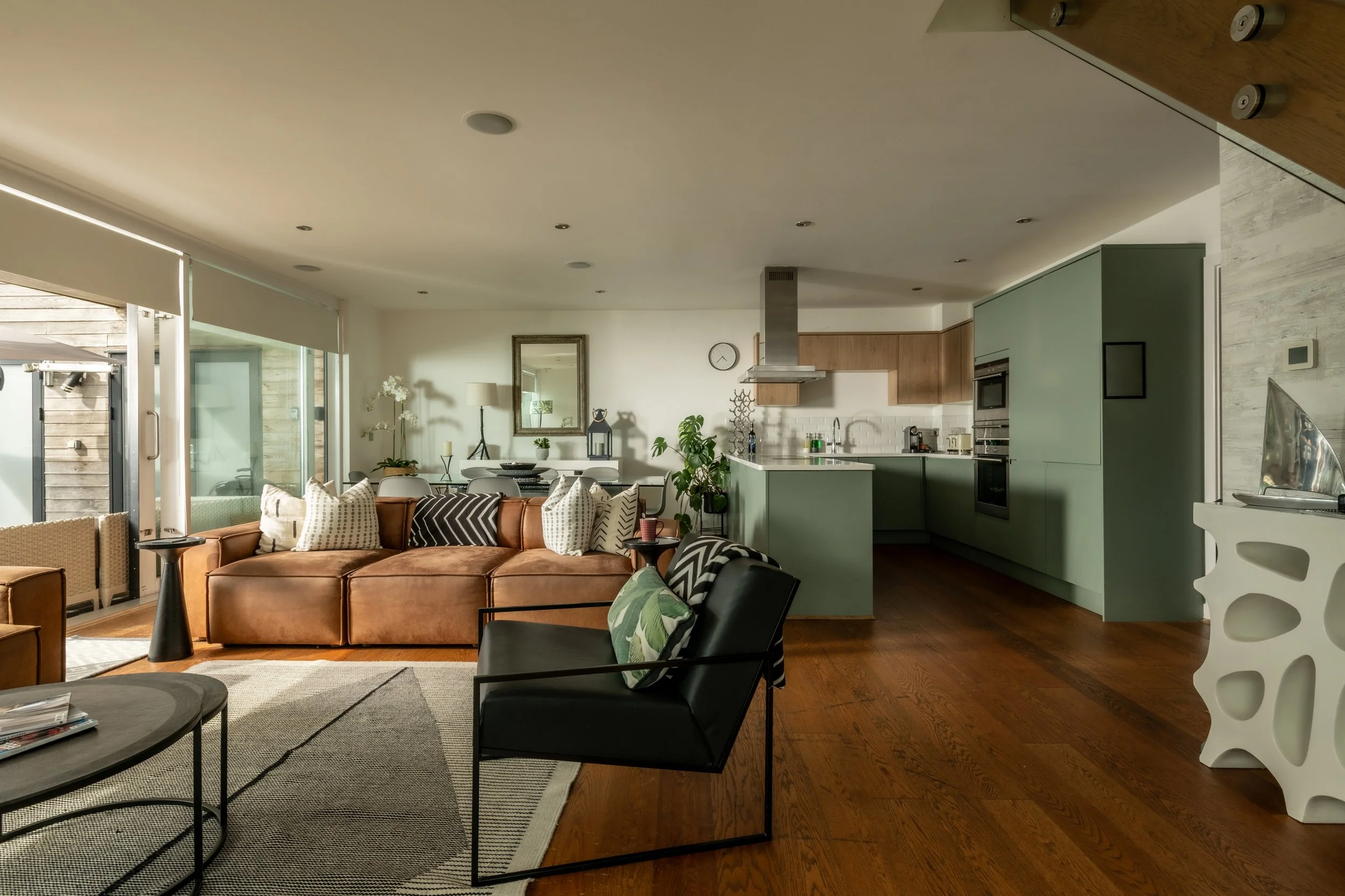 Open-concept living room and kitchen with brown leather sofa, black chairs, wooden flooring, green kitchen cabinets, and large windows letting in natural light.