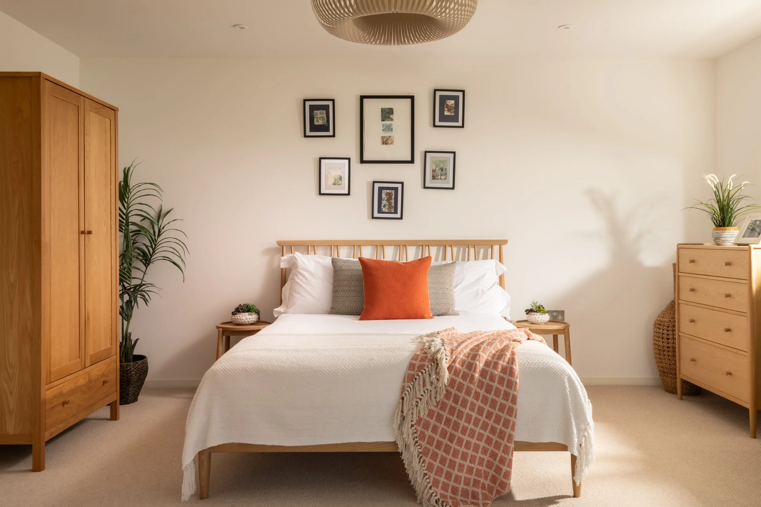 A cozy bedroom with a minimalist design featuring a wooden bed with white bedding and decorative pillows, a wooden wardrobe on the left, a small wooden nightstand with plants, and a light-colored dresser on the right. The wall above the bed has a gal
