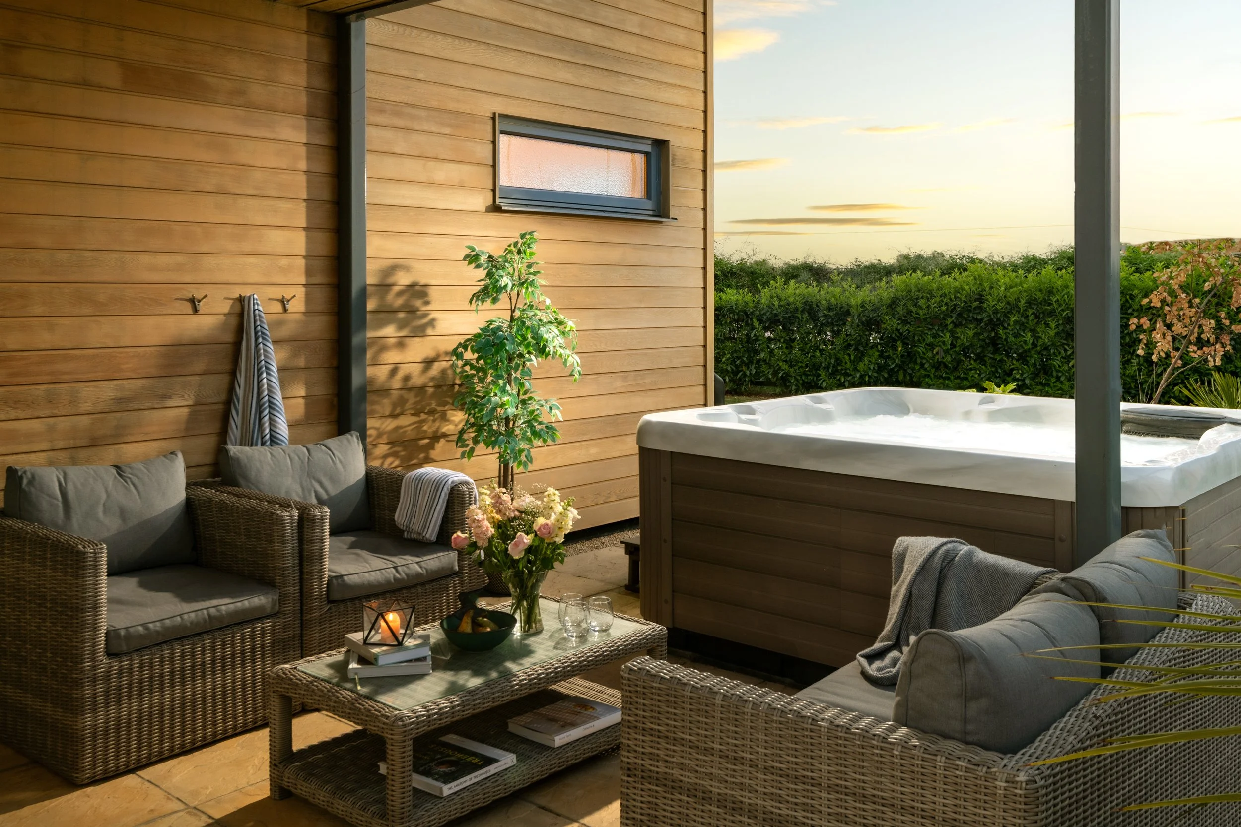 Outdoor patio with wicker furniture, a glass-topped coffee table, and an outdoor hot tub, set against a wooden wall with a small window, greenery, and a sky with clouds at sunset.