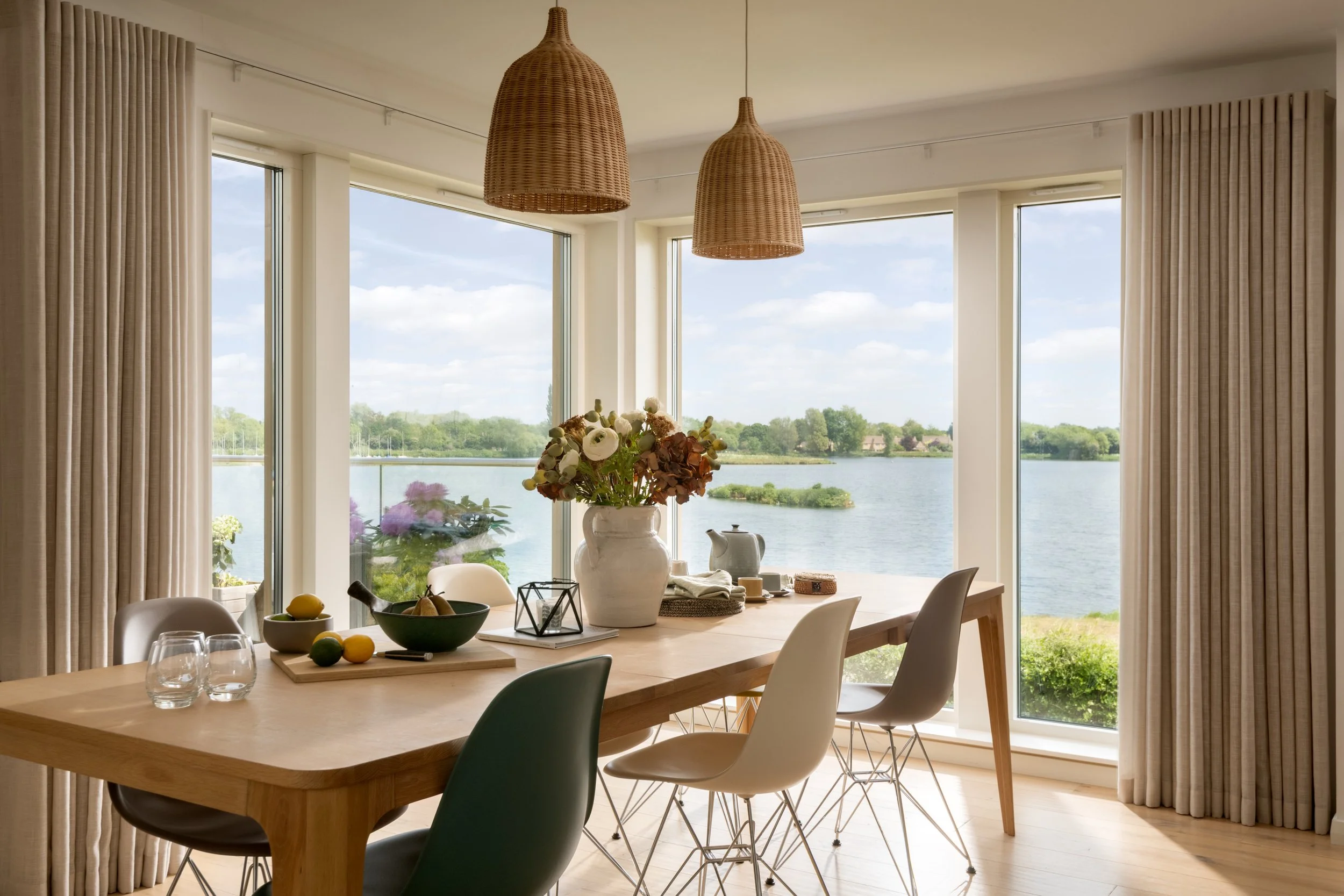 A dining room with a wooden table, white and green chairs, and a large window overlooking a river with trees in the background, decorated with a vase of flowers and fruits on the table, and wicker pendant lights hanging from the ceiling.