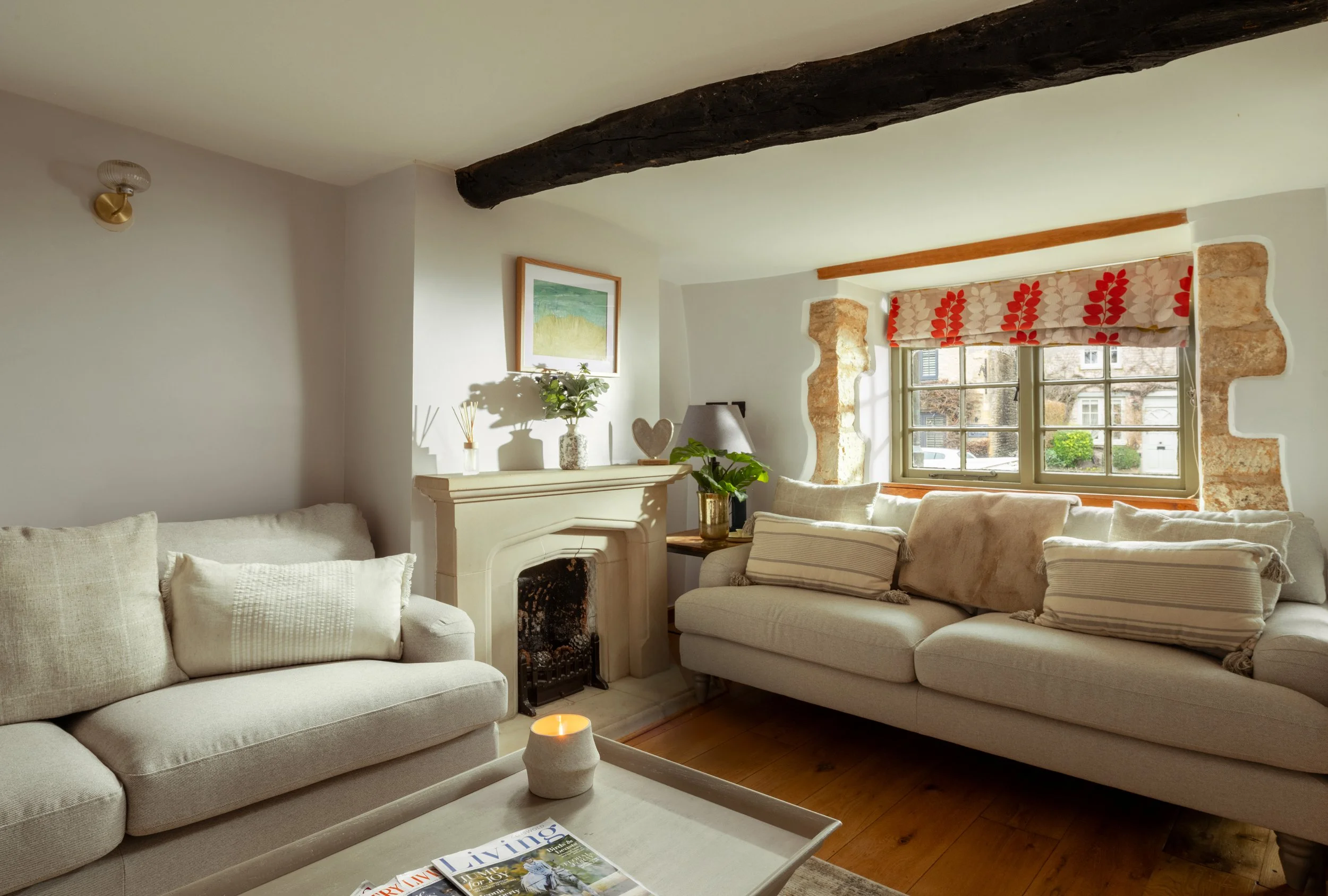 Living room with cream-colored sofa, armchair, fireplace, framed picture, potted plant, window with patterned curtain, and wooden beam ceiling.